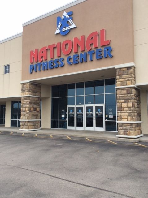 Entrance to a National Fitness Center with a large red and blue sign, stone pillars, and glass doors.