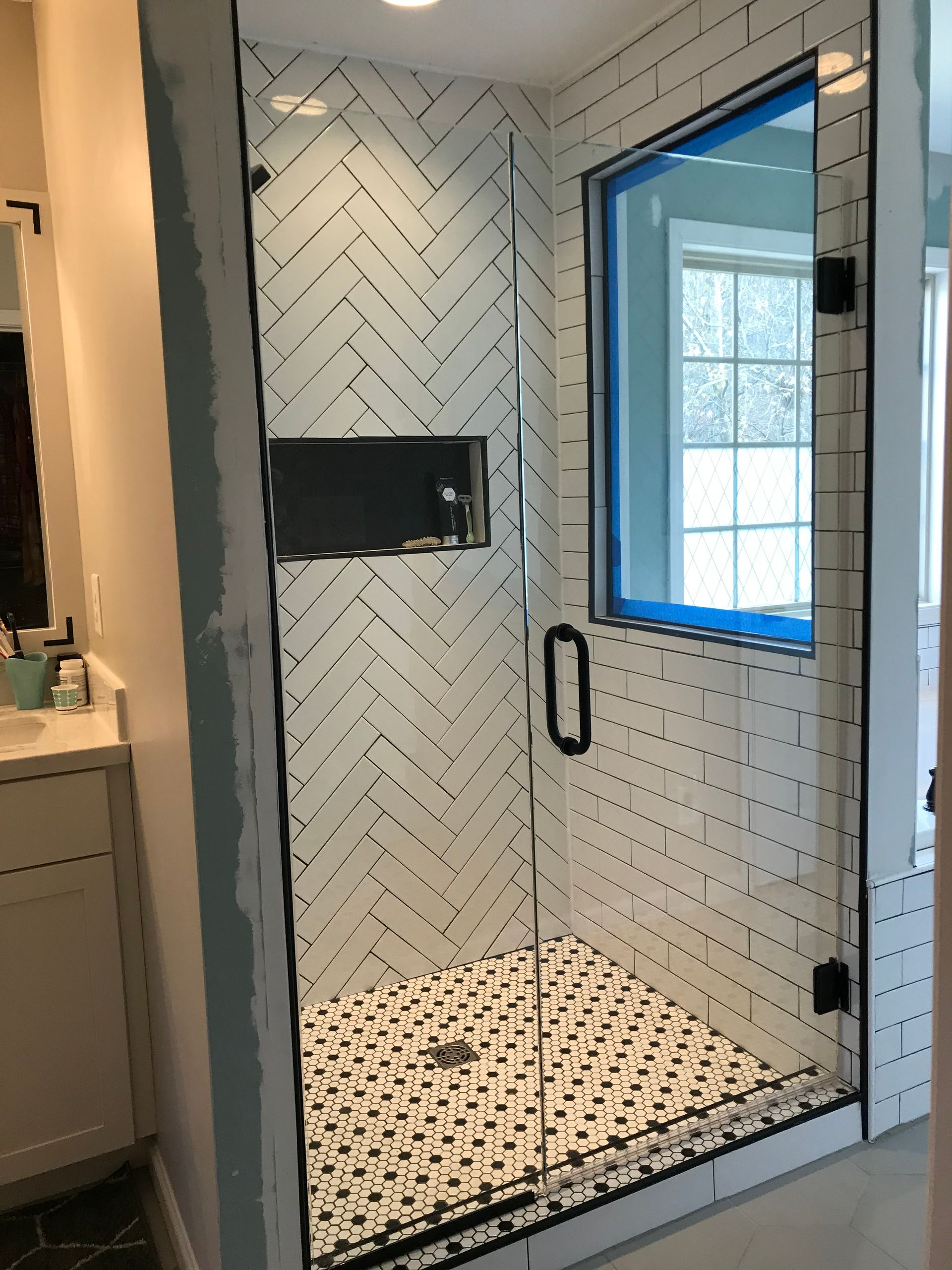 A walk-in shower with white herringbone tile walls, a black niche, a patterned tile floor, and a glass door.