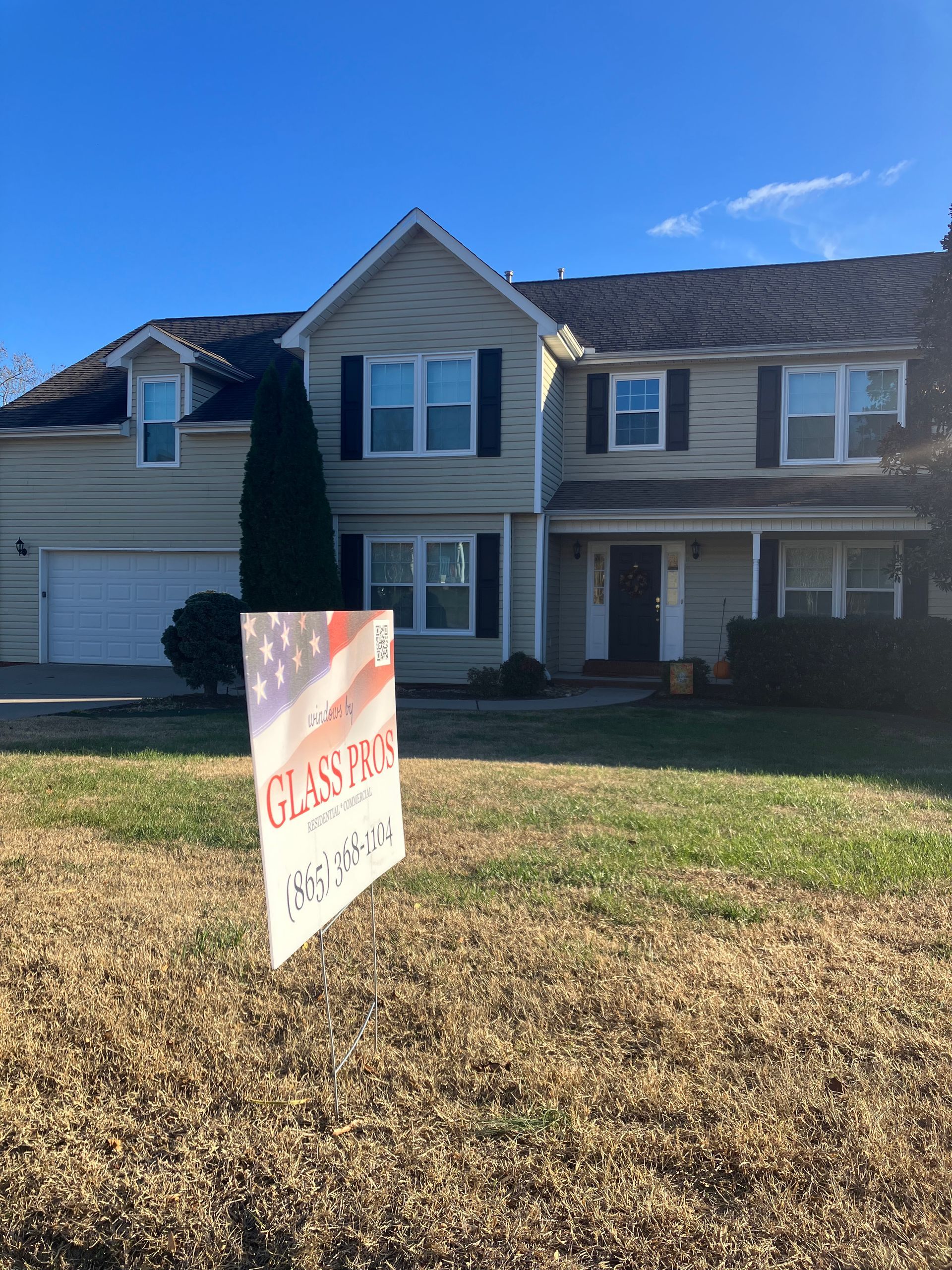 A tan two-story suburban house under a clear blue sky, with a promotional sign in the foreground lawn.