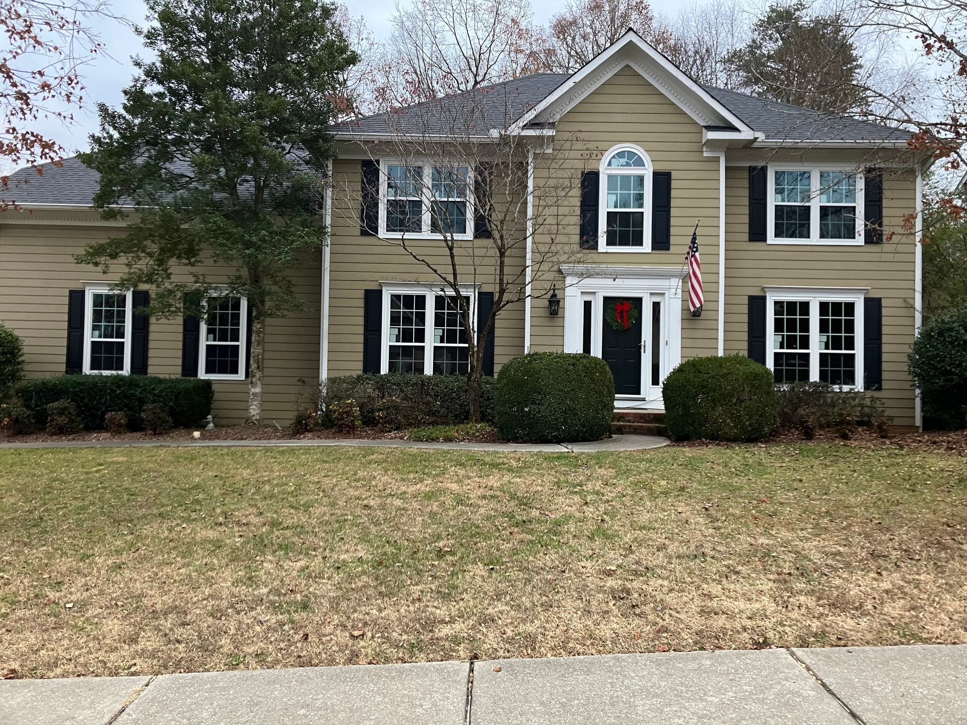 Two-story suburban tan house with black shutters, a gabled roof, a central front door, and a manicured lawn.