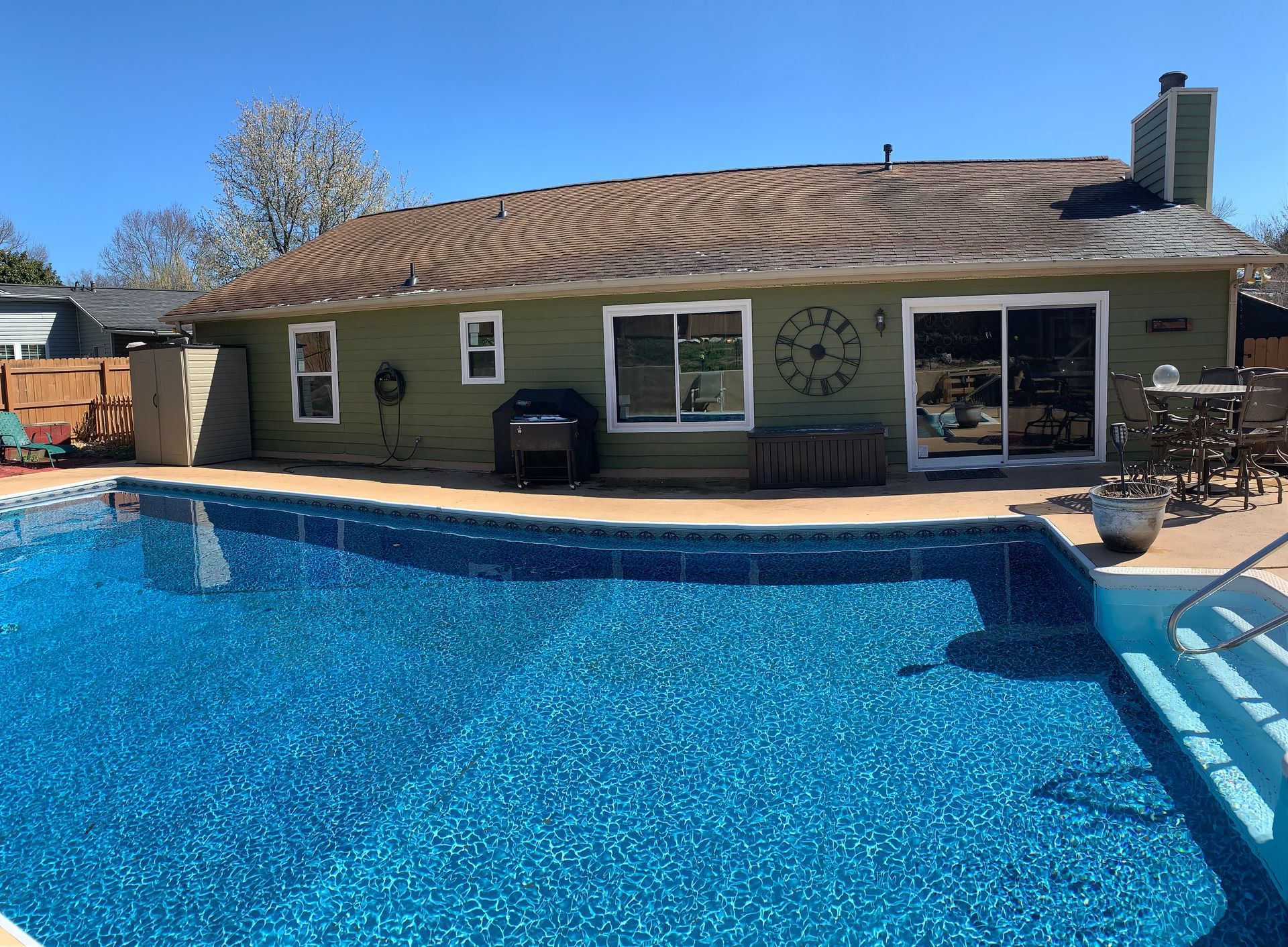 A backyard swimming pool with blue water sits in front of a green, single-story house with a patio and sliding doors.