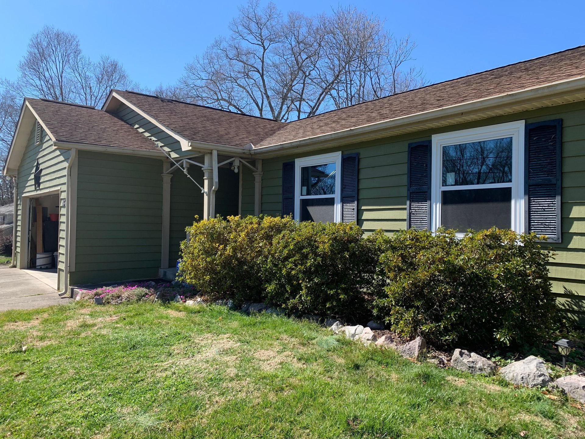 A green one-story suburban house with dark shutters, a garage, and a landscaped front yard under a clear blue sky.