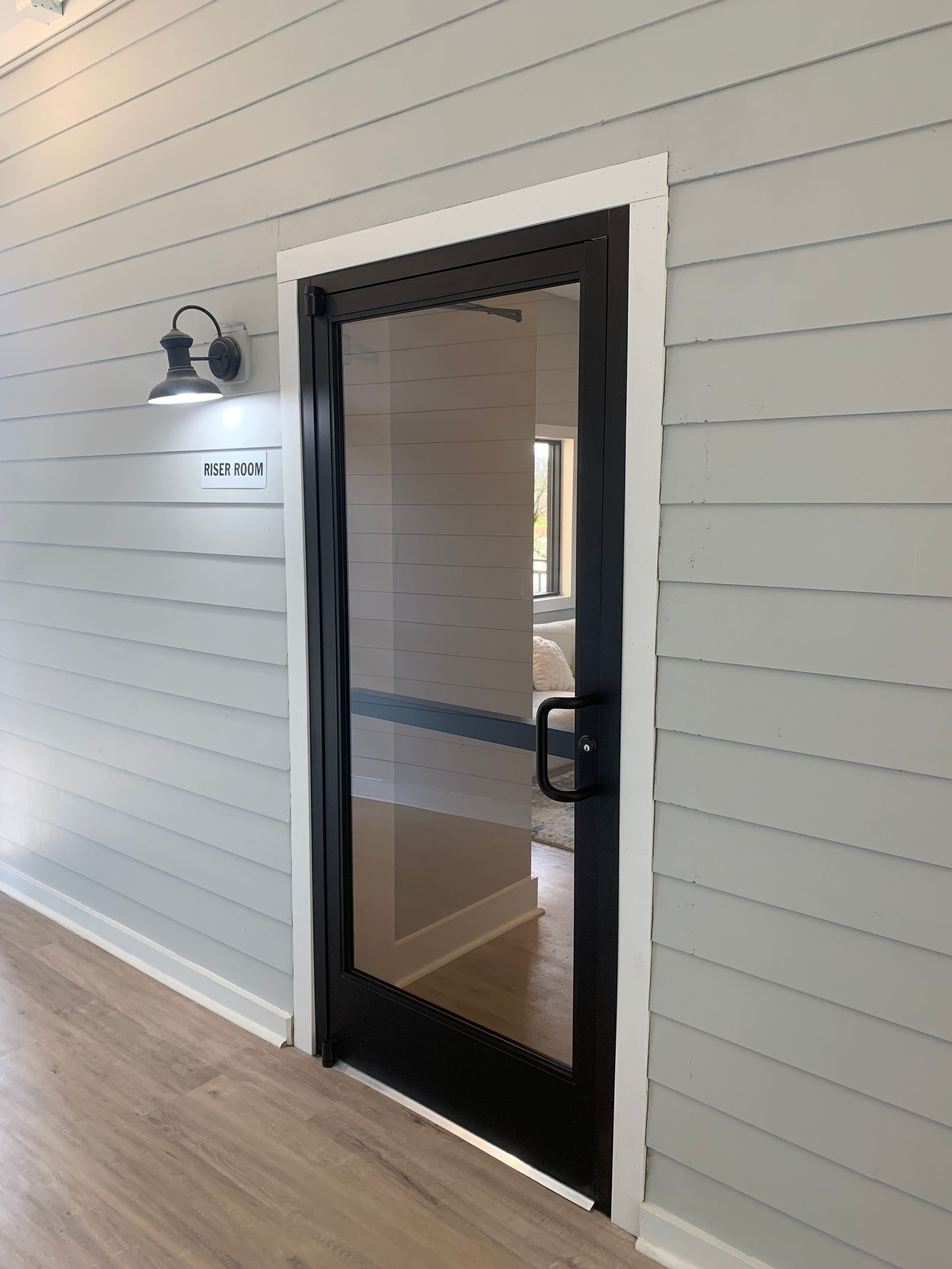 A black-framed glass door with a handle, set in a light gray horizontal shiplap wall next to a wall-mounted light fixture.