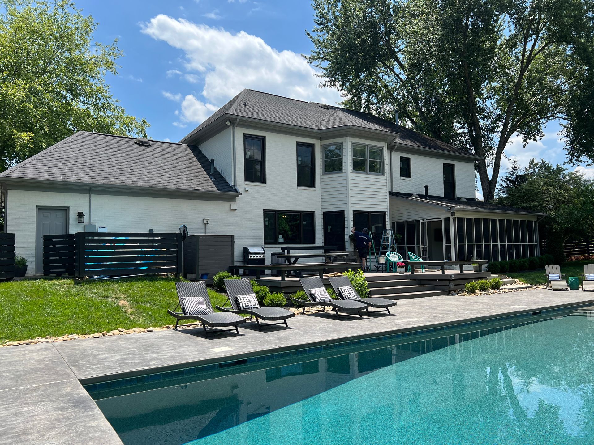 A white two-story house with a grey roof and a stone patio surrounding a swimming pool with lounge chairs.