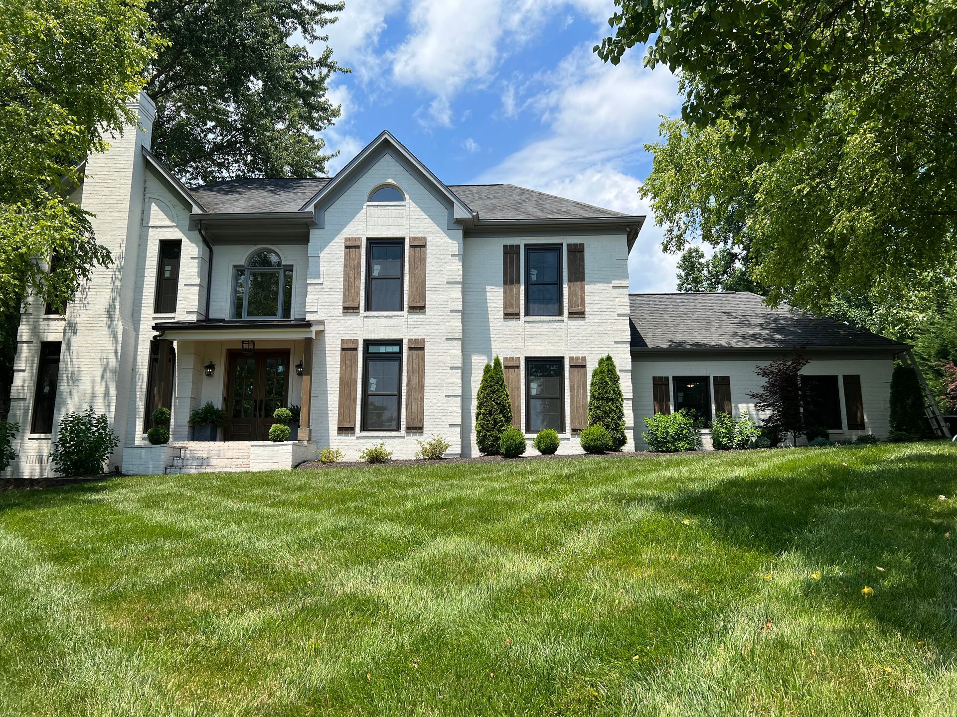 A large, two-story white brick house with a dark shingled roof, set back from a manicured green lawn under a blue sky.