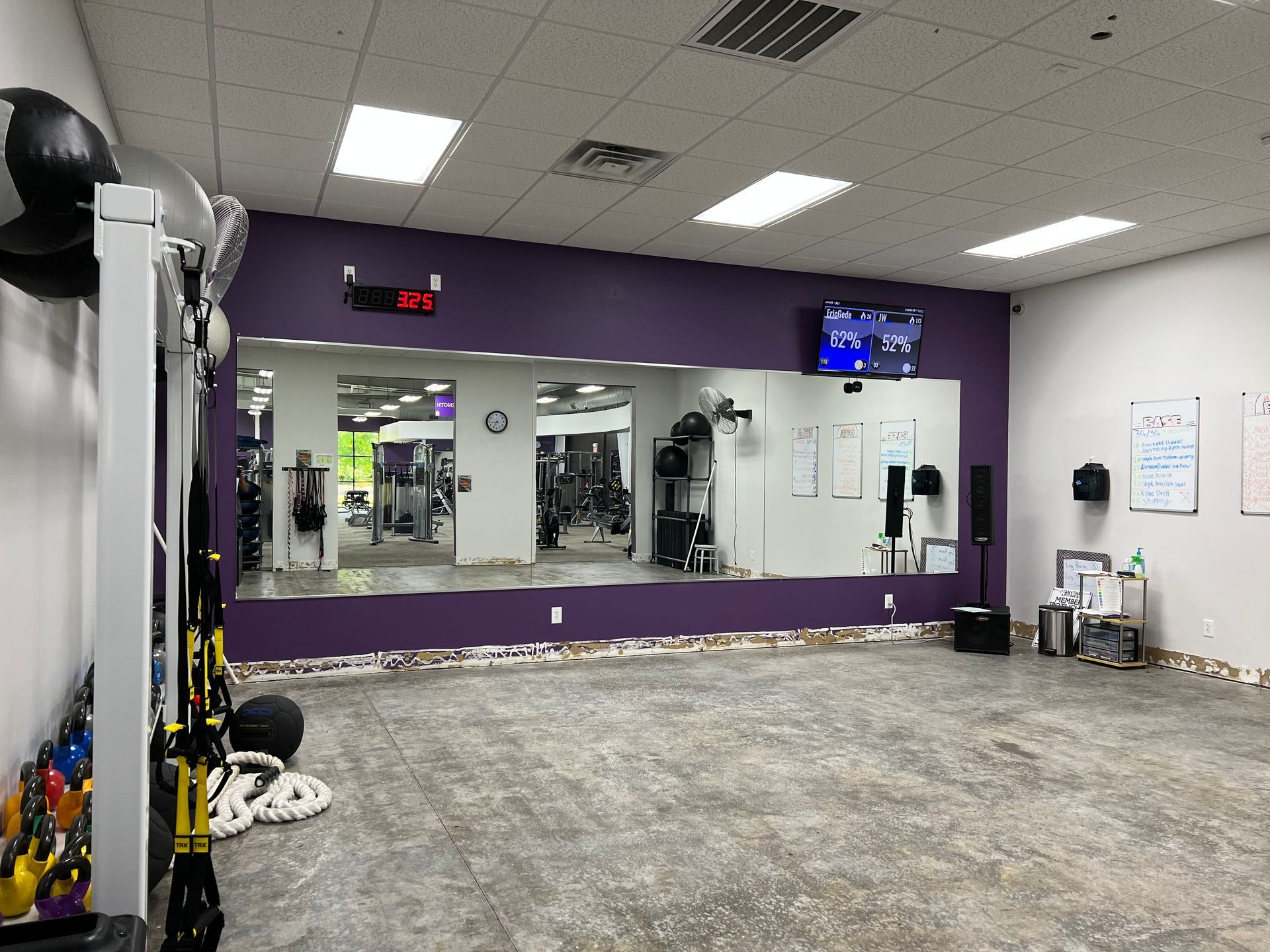 Empty fitness studio with a purple wall, large mirrors, a flat-screen TV, and exercise equipment in the foreground.