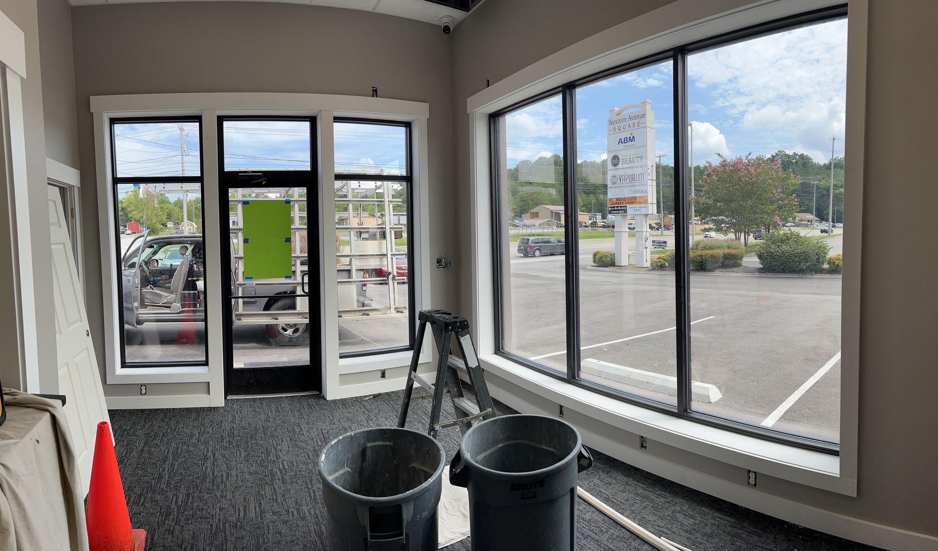 An interior view of a room under construction with large windows, two gray trash cans, and a ladder on grey carpet.