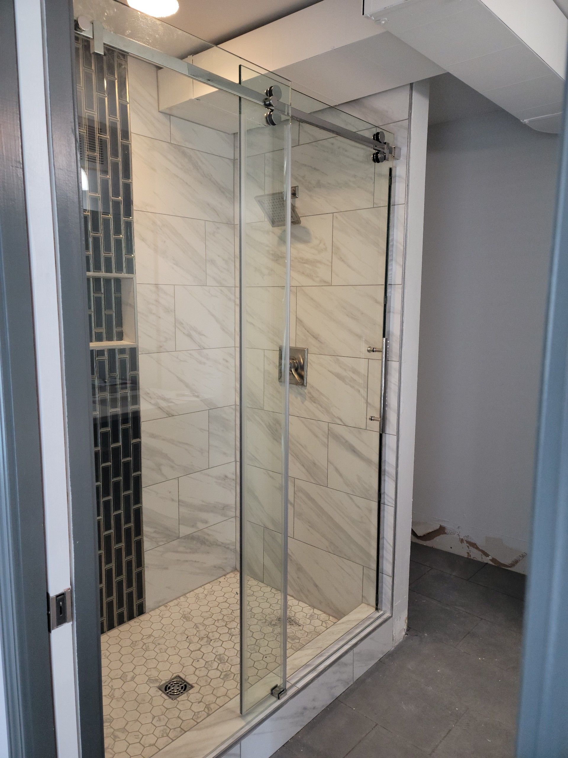 A walk-in shower with a glass sliding door, marble-look tiled walls, a dark decorative accent strip, and stone floor tiles.