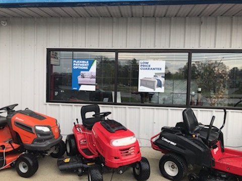 Three lawn mowers parked in front of a store window with signs for flexible payment options and a low price guarantee.