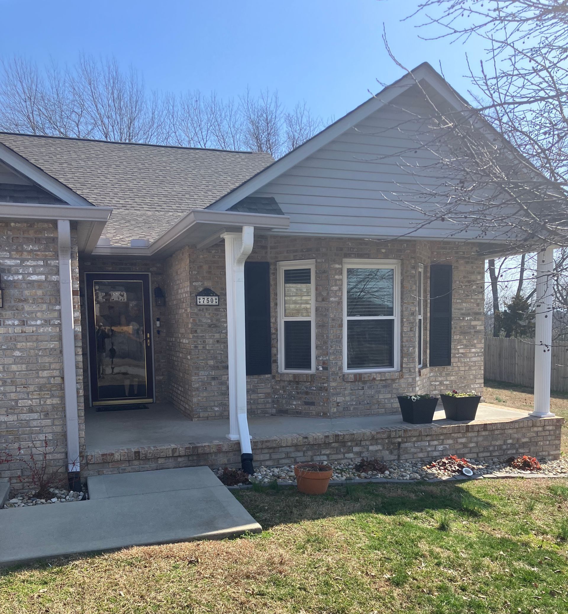 A tan brick house with a front porch, grey shingles, and white pillars under a clear blue sky on a sunny day.
