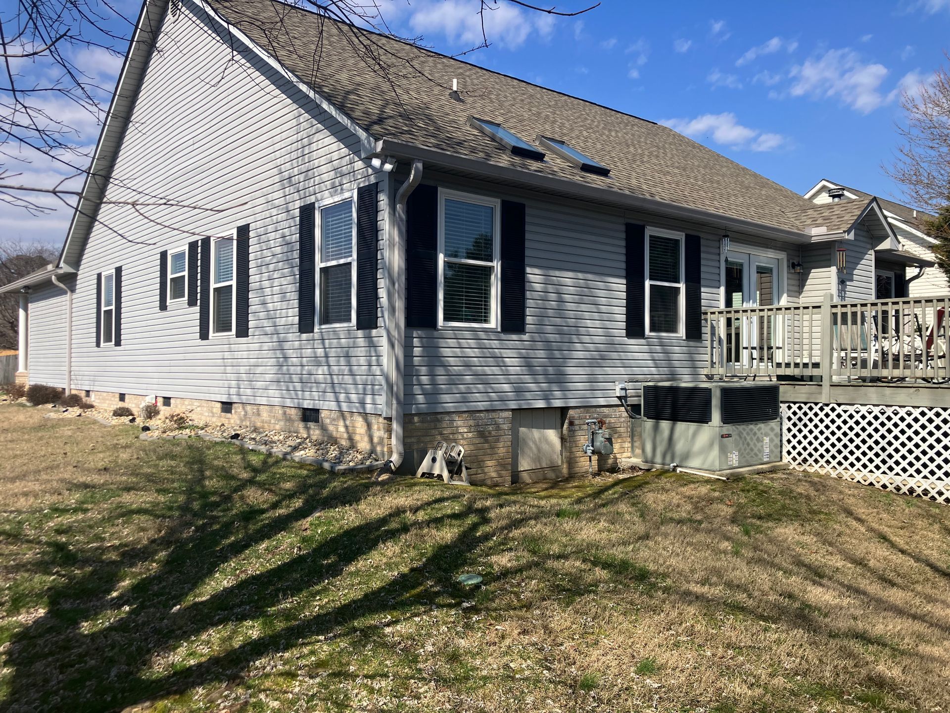 A gray, single-story house with a shingled roof, side windows, a deck, and an HVAC unit on a grassy slope.