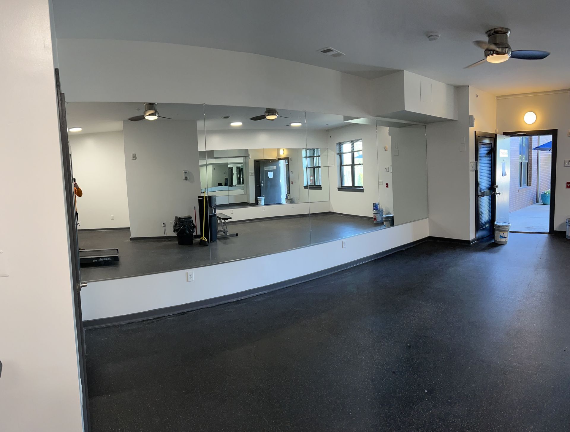 A bright, empty fitness studio featuring dark rubber flooring, floor-to-ceiling mirrors, ceiling fans, and an open doorway.