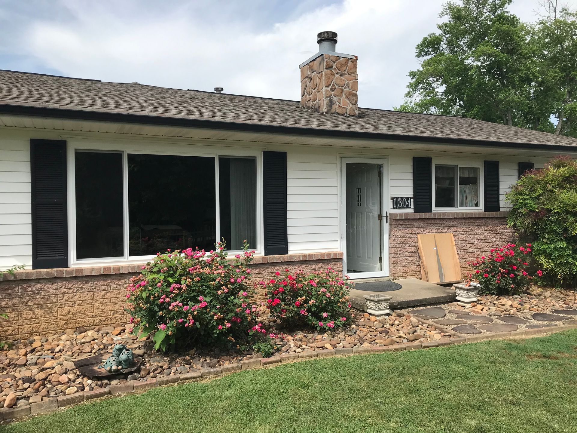 A single-story ranch-style house with white horizontal siding, black shutters, a brick facade, and a stone chimney.