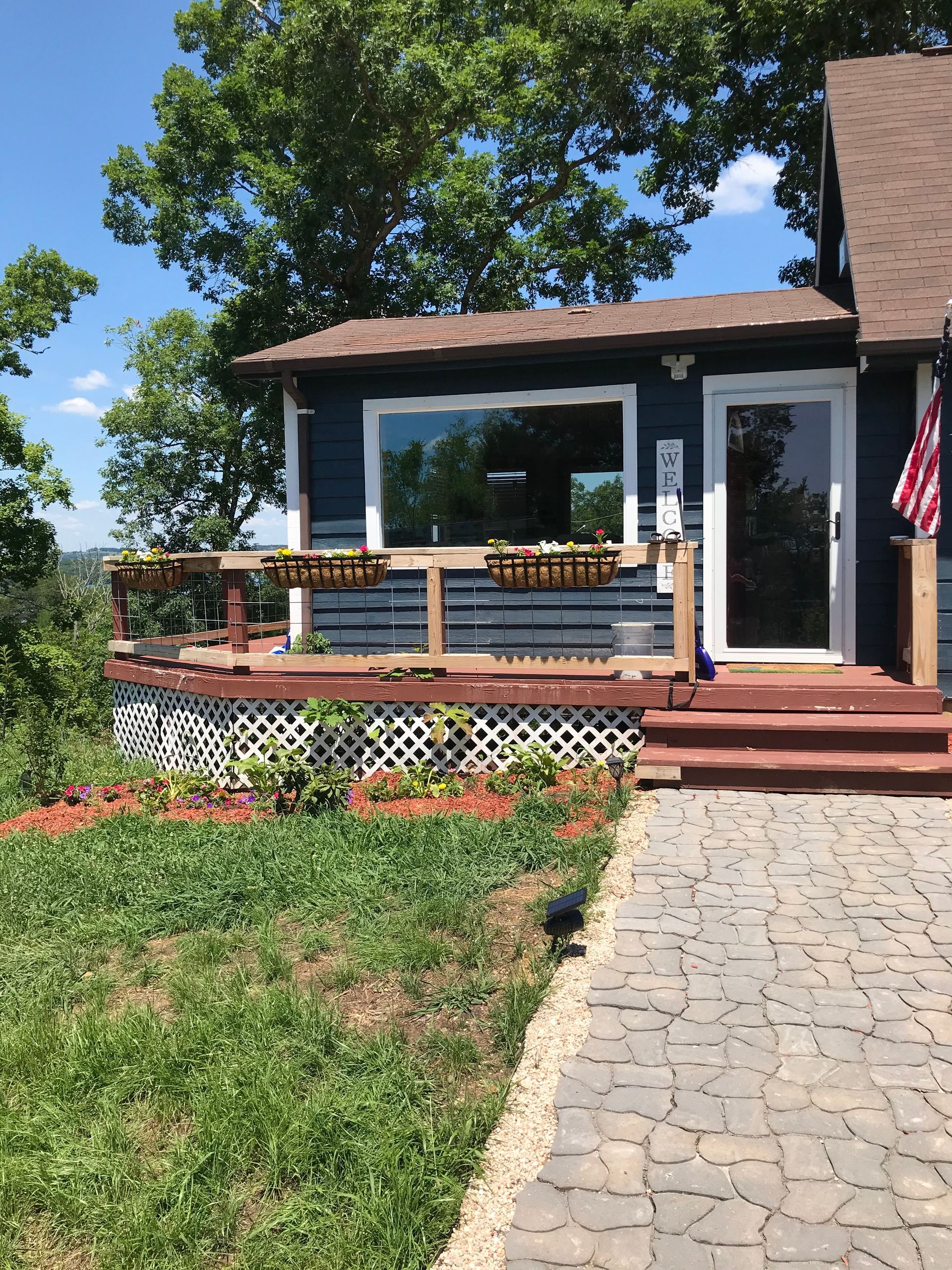 A blue house with a wooden deck, lattice skirting, and a stone walkway in a sunny, tree-filled setting.