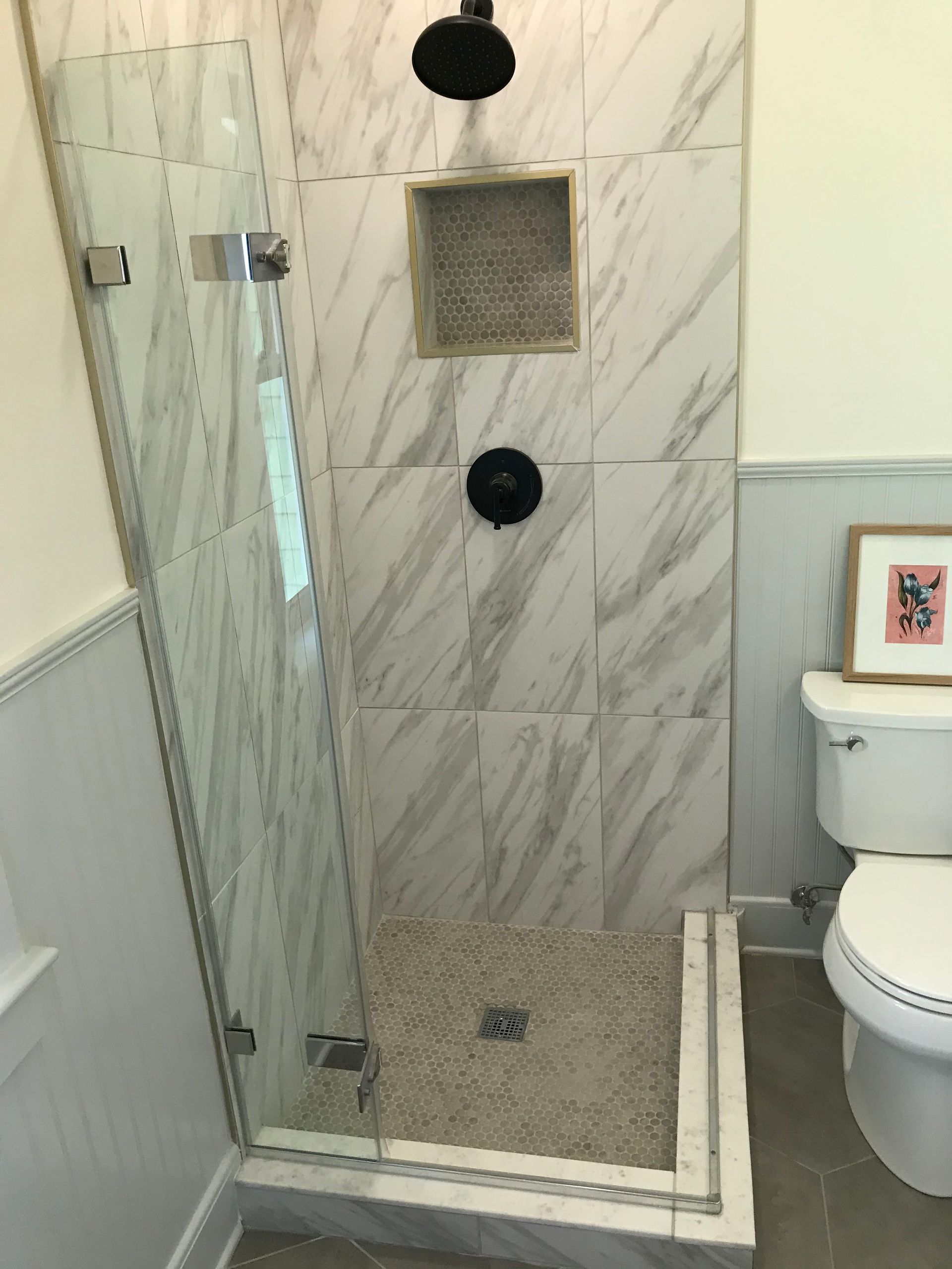 A walk-in shower with marble-look tiled walls, pebble floor, glass door, black fixtures, and a wall niche beside a toilet.