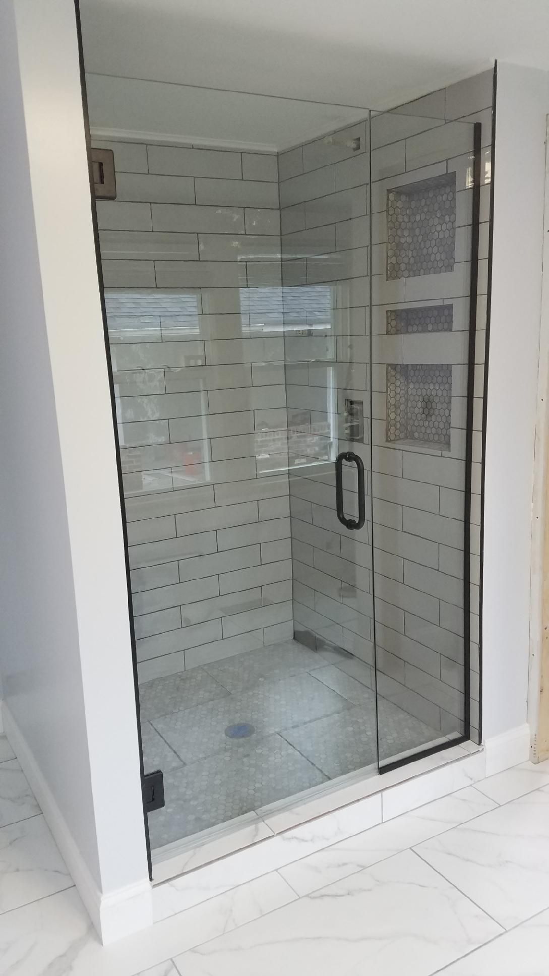 A glass-enclosed shower with white subway-style wall tiles and gray floor tiles, featuring a black handle and door hinges.