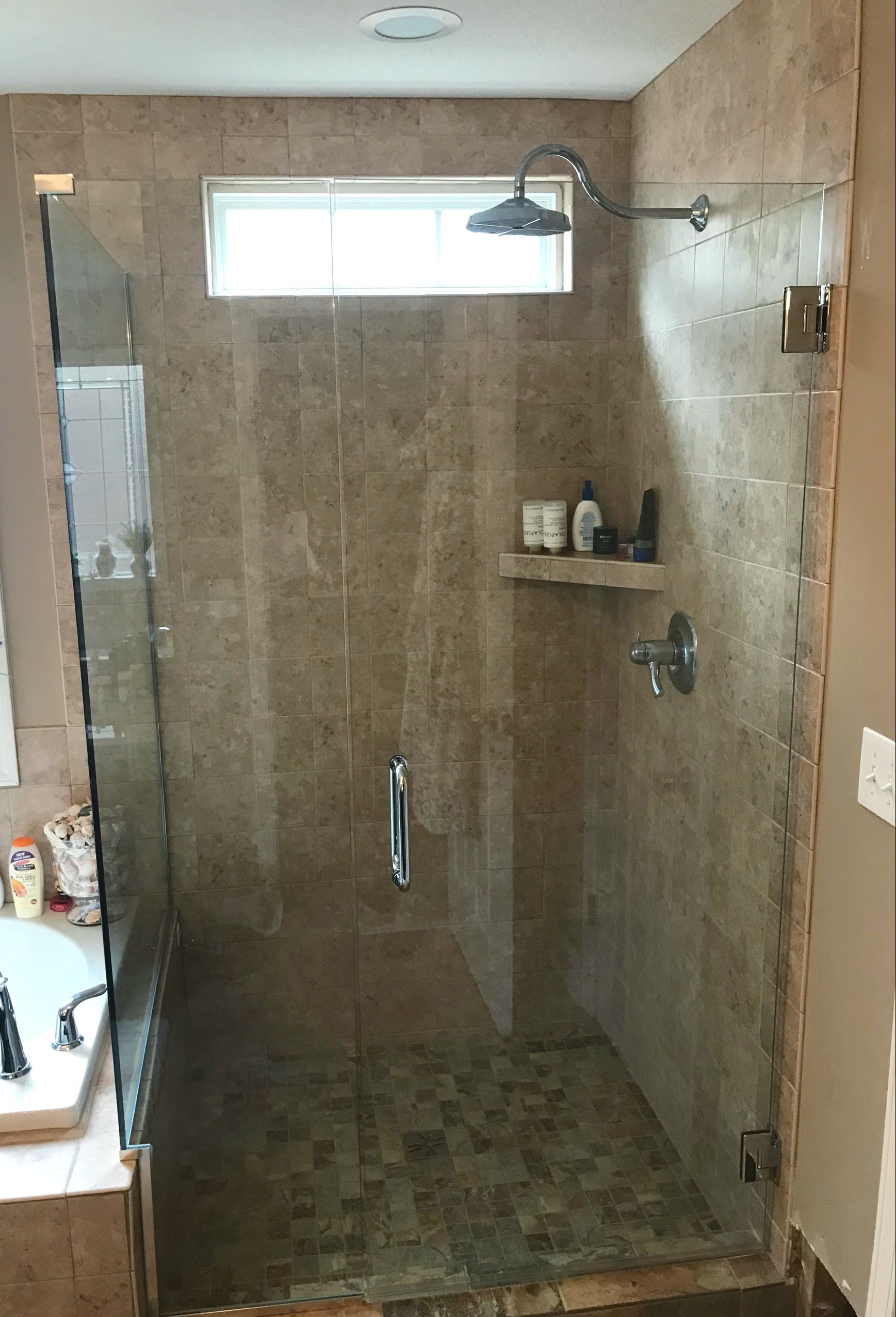 A glass-enclosed walk-in shower with brown tiled walls and floors, featuring a mounted showerhead and a corner shelf.