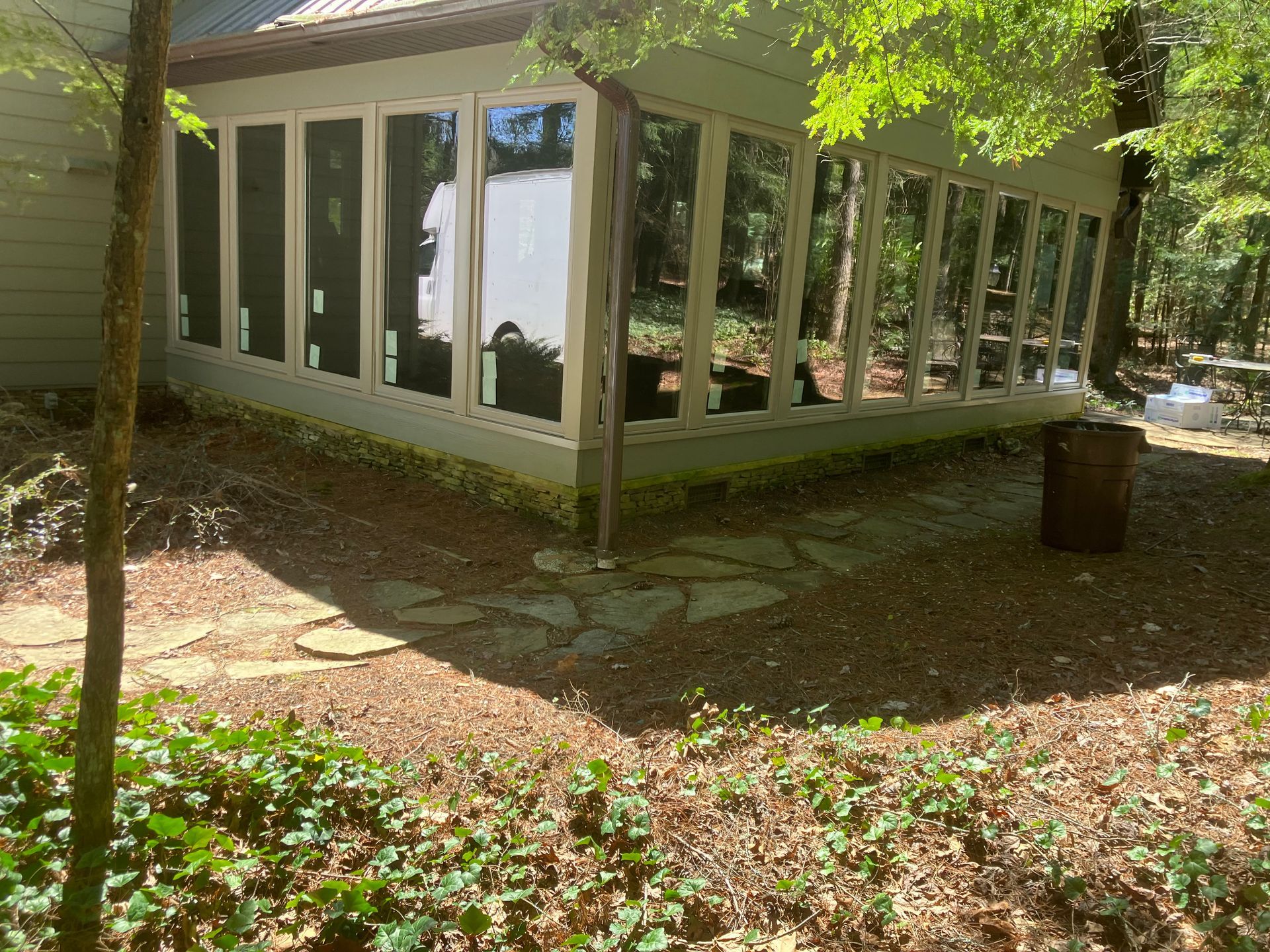 A sunroom with large windows and white trim sits in a wooded yard with a stone path and a metal barrel.