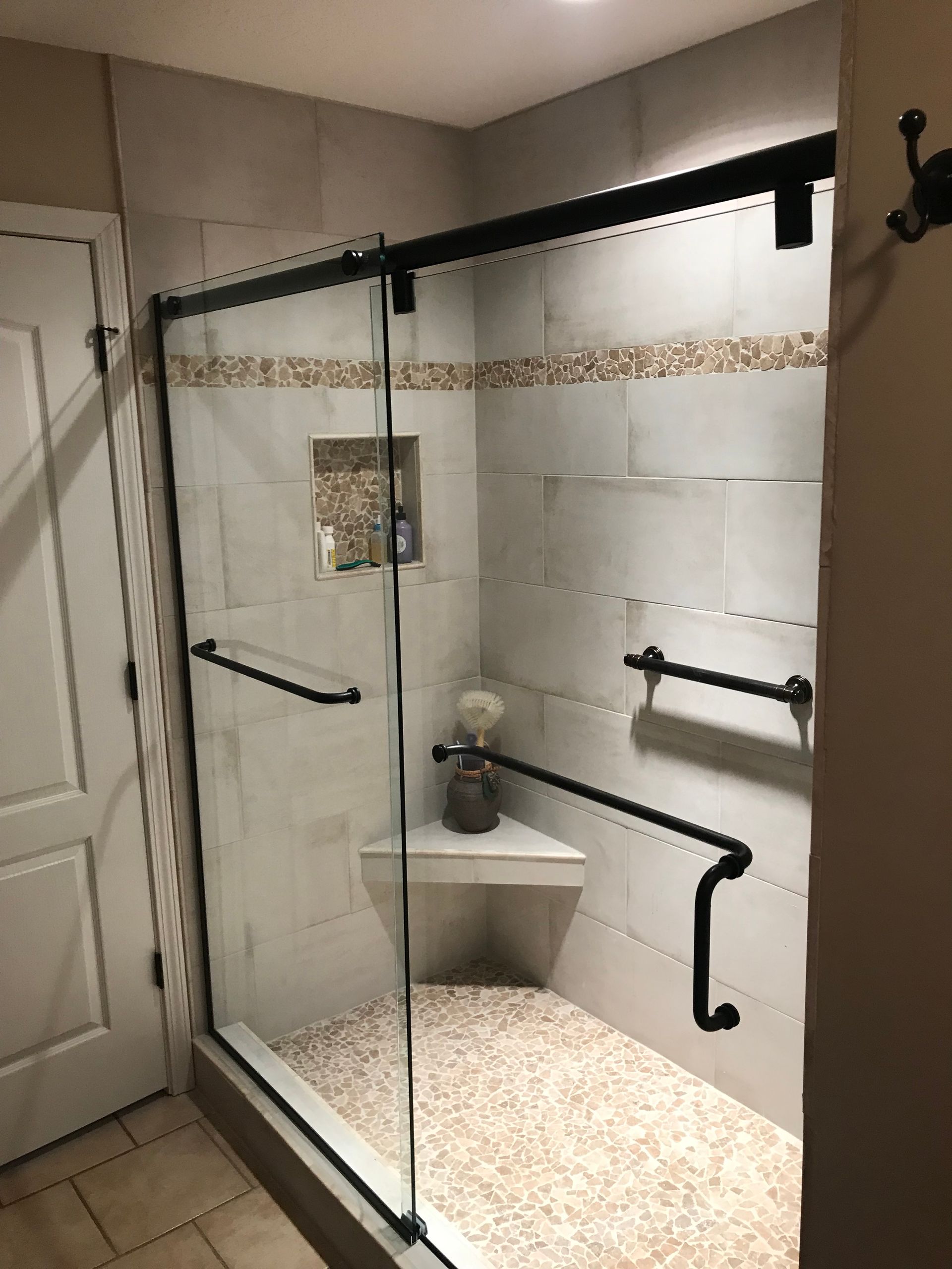 A glass-enclosed shower with tiled walls, a corner bench, a built-in niche, and black grab bars and door hardware.