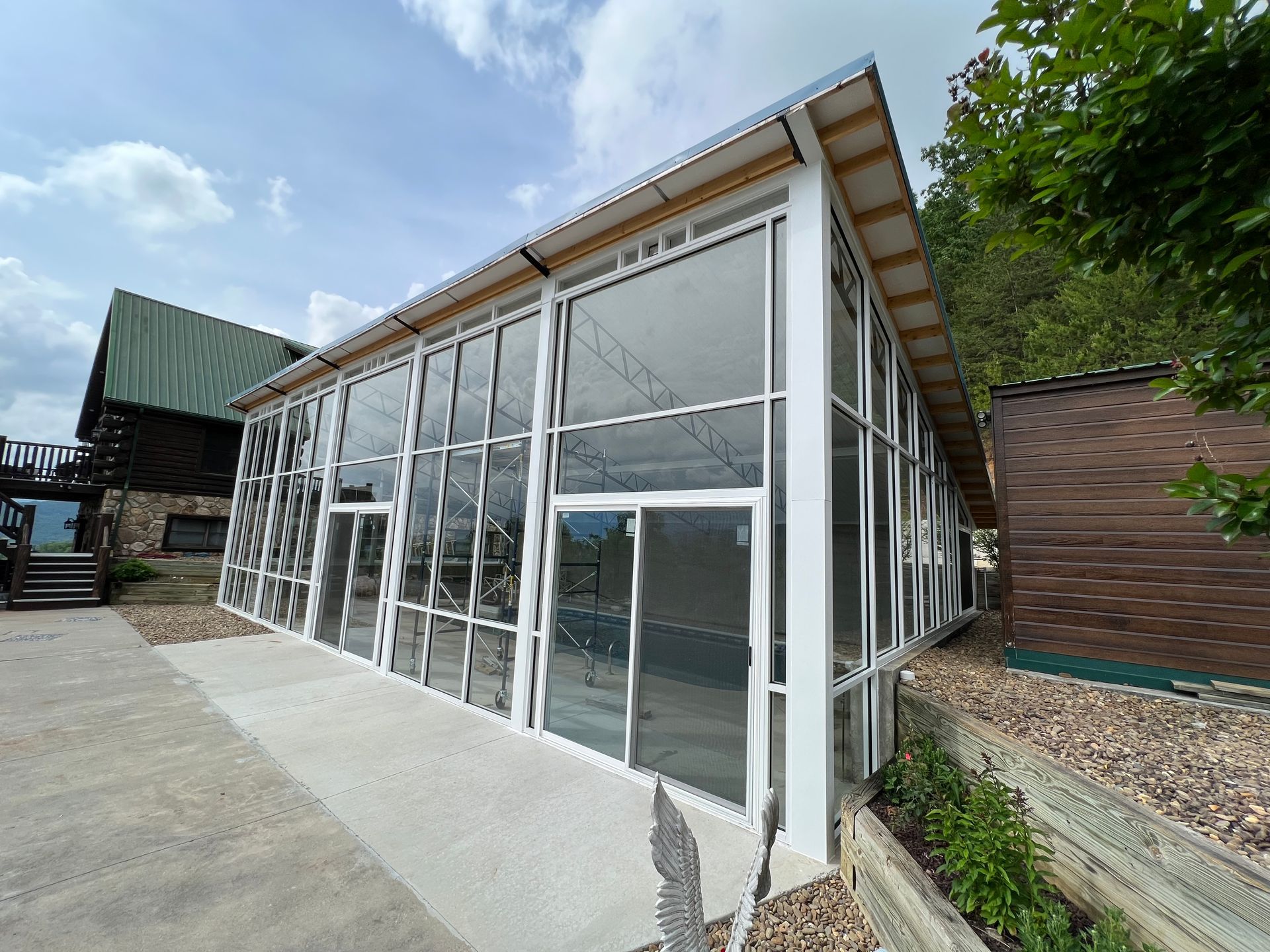 A modern, white-framed glass sunroom or conservatory with a sloped roof stands on a concrete patio next to wooden buildings.