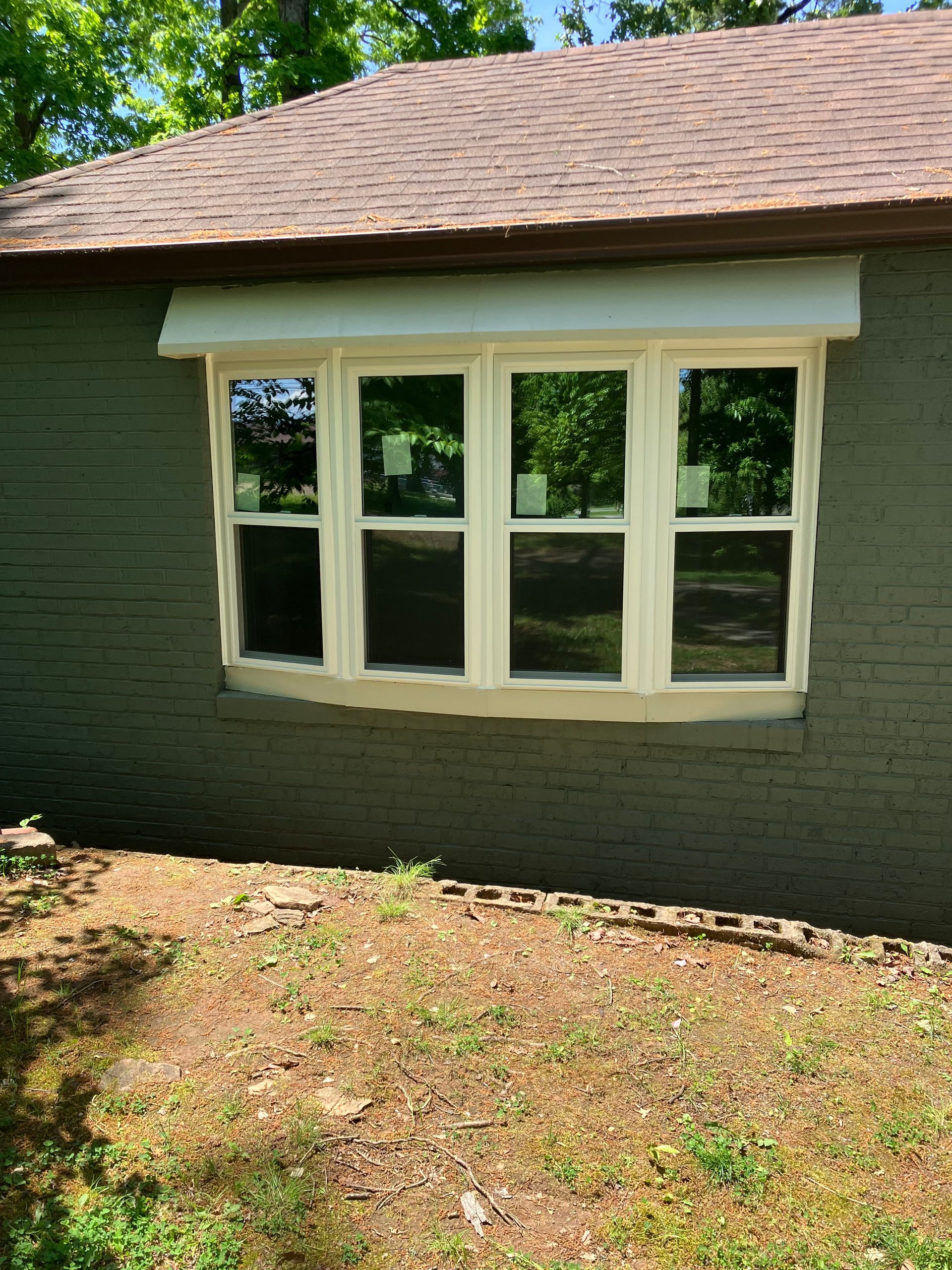 A white, four-pane bay window on a green brick exterior wall of a house with a brown shingled roof and mulch landscaping.
