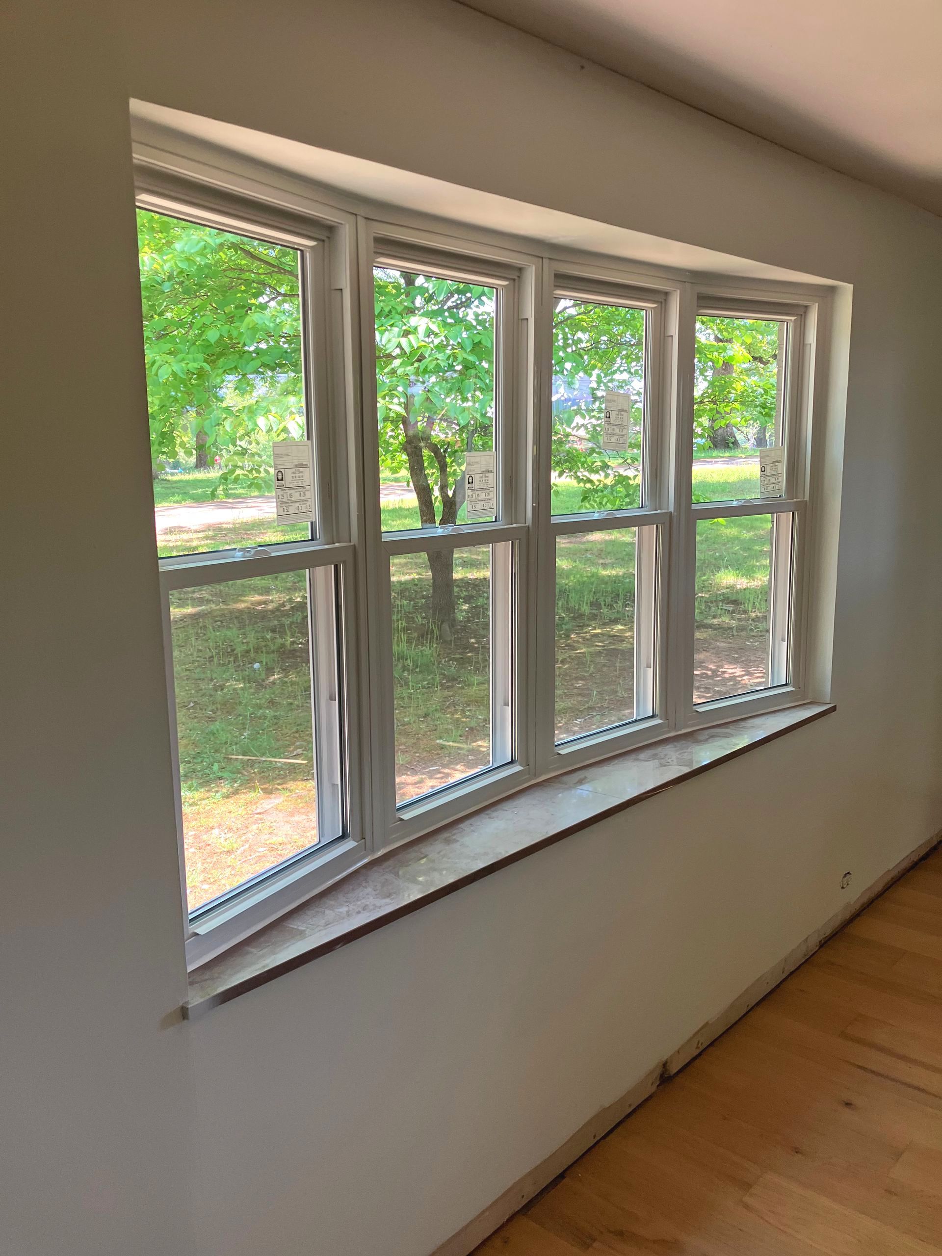 A wide, white-framed window with four panels looks out onto a green, tree-filled yard from a room with light wood floors.