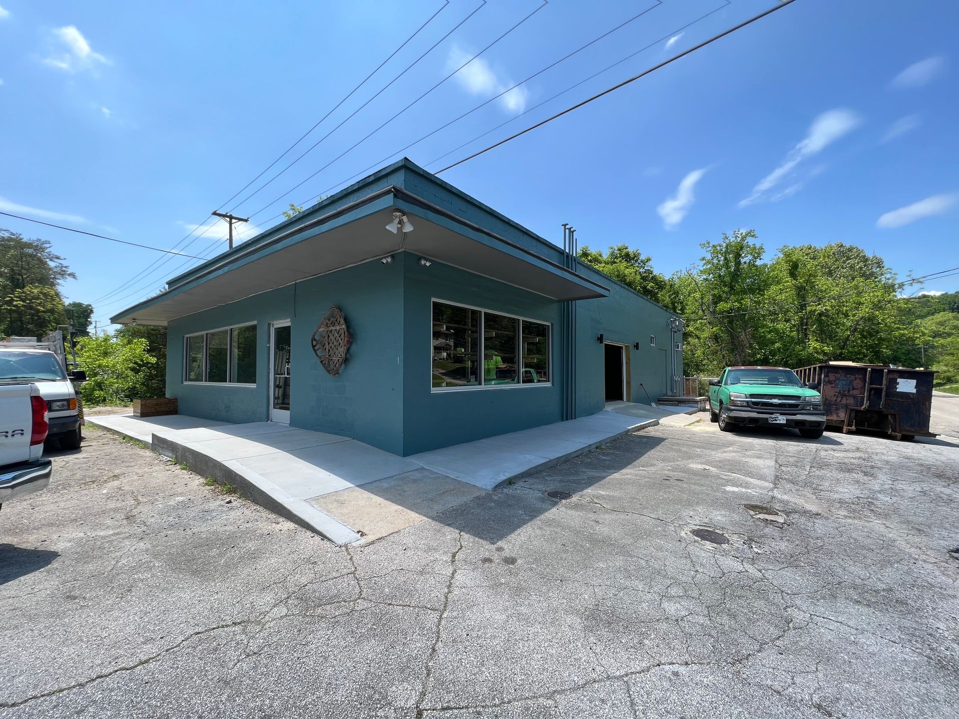 A teal, flat-roofed commercial building with a ramp entrance, surrounded by a gravel parking lot under a sunny blue sky.