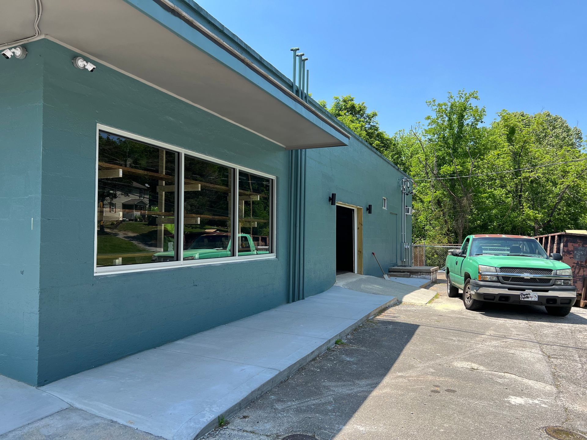 A teal building exterior featuring a large window, a covered entrance, and a green pickup truck parked on the pavement.