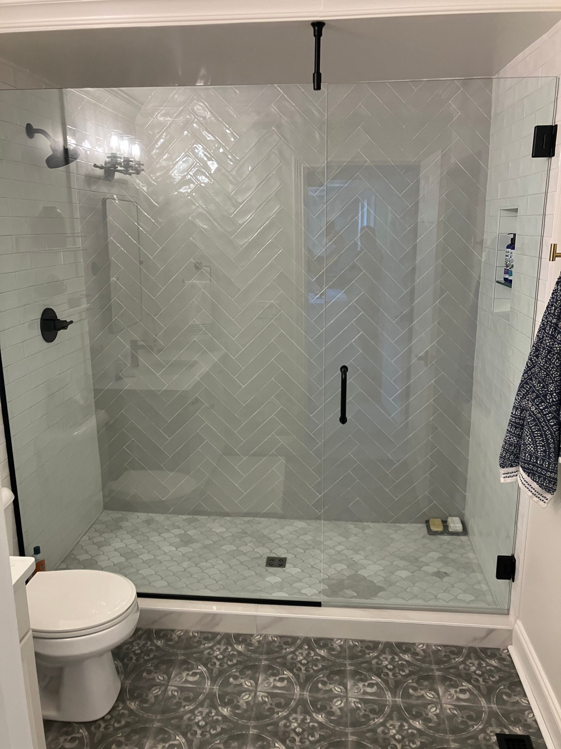 A modern bathroom featuring a glass-enclosed shower with white herringbone-patterned tile walls and a patterned floor.