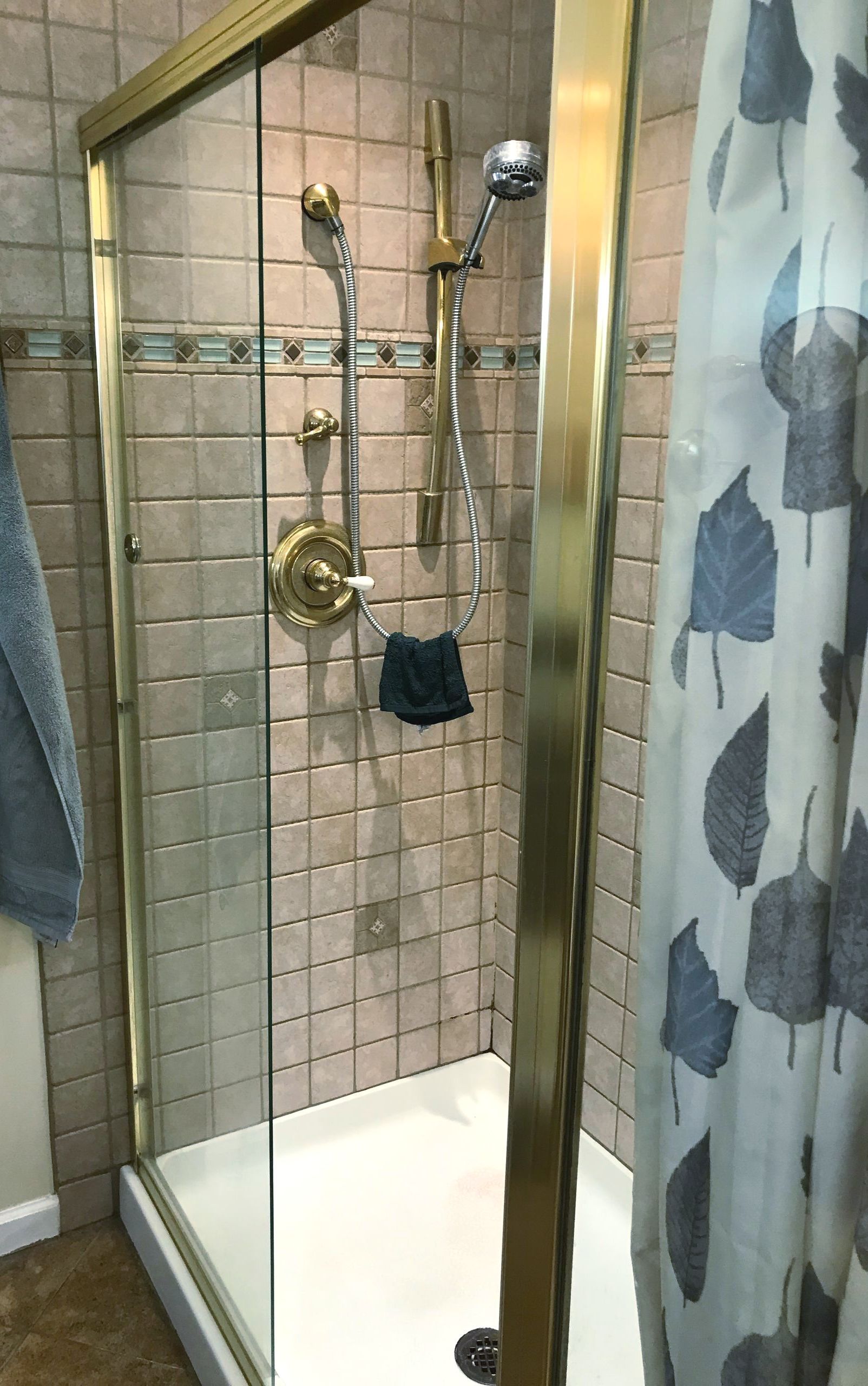 A shower stall with tan tiled walls, brass trim, a sliding glass door, and a handheld shower head on a vertical bar.