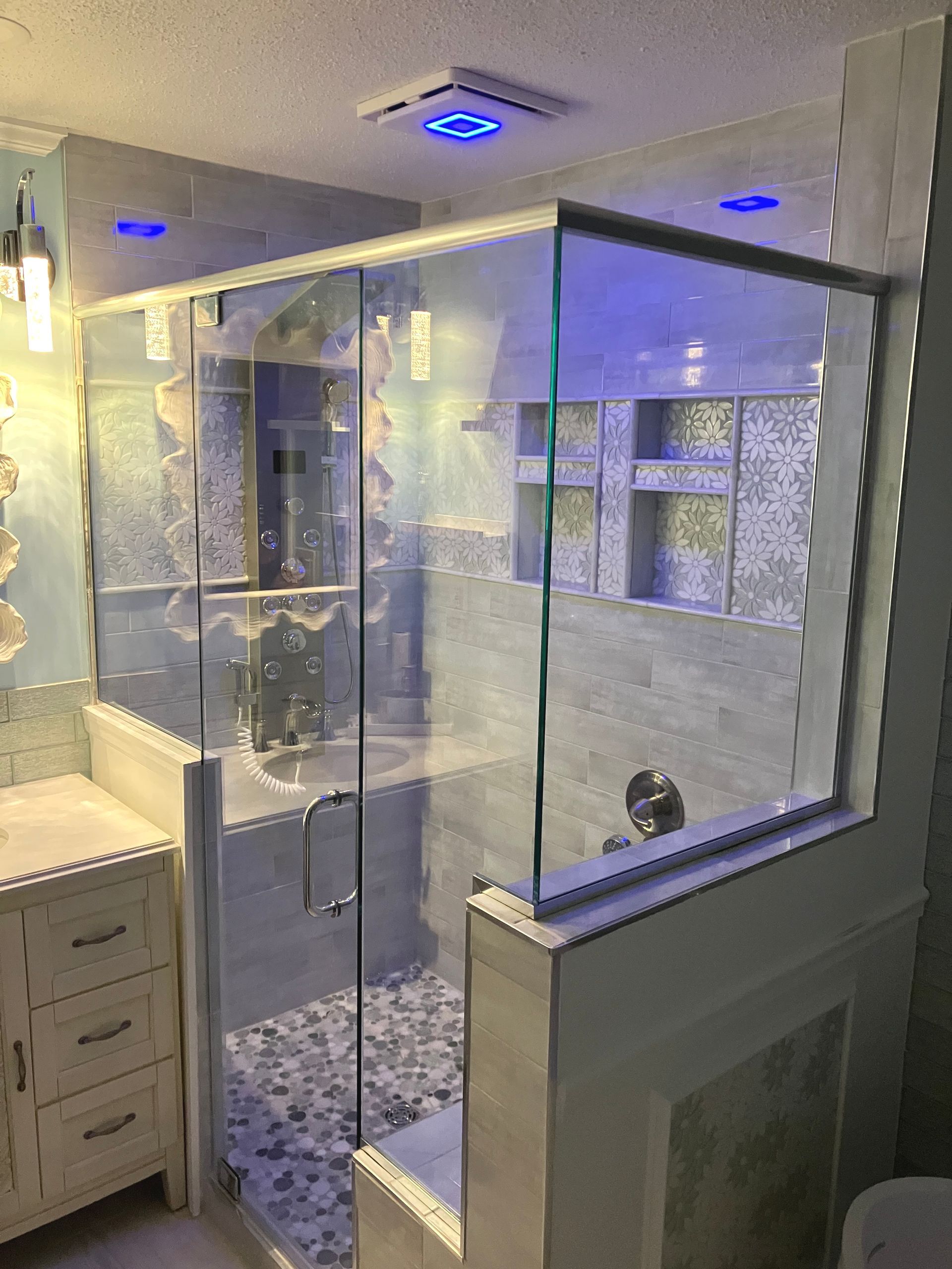 A modern bathroom with a glass shower enclosure, blue LED ceiling lights, and patterned tile accents.