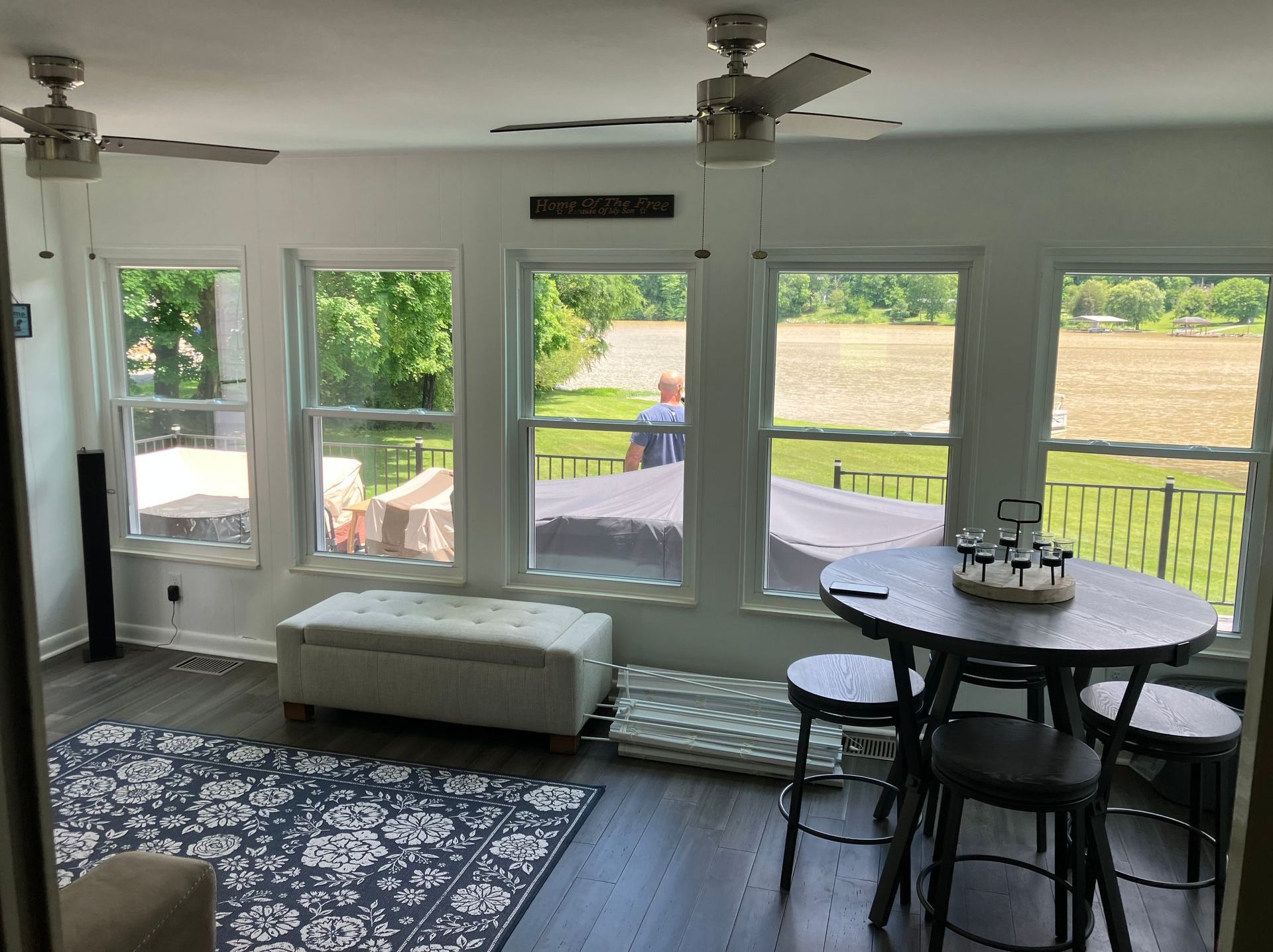 A sunroom with dark wood floors, large windows overlooking a field, two ceiling fans, a dining set, and an ottoman.
