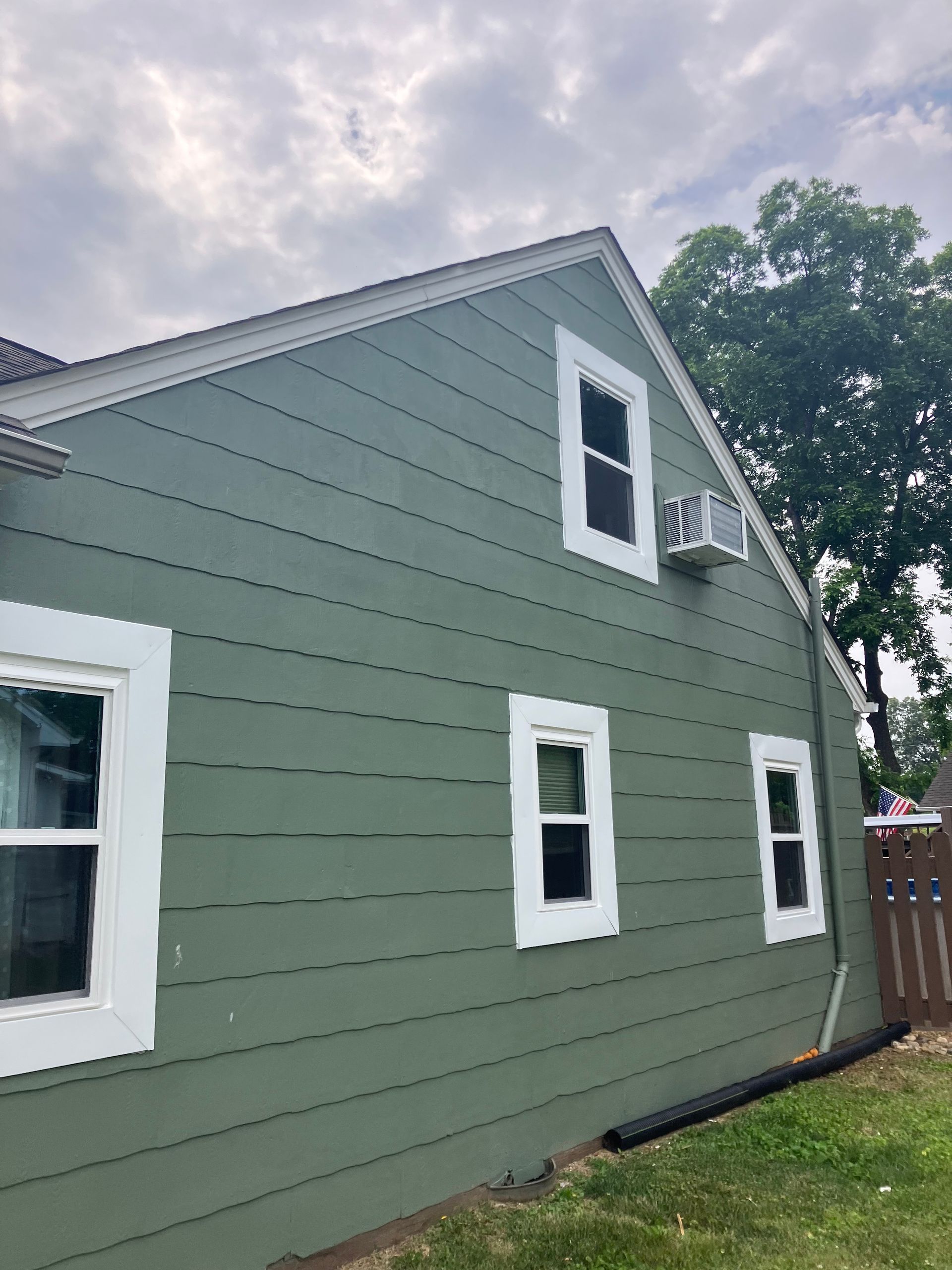A side view of a sage green house with white window trim and an air conditioning unit installed in an upper window.