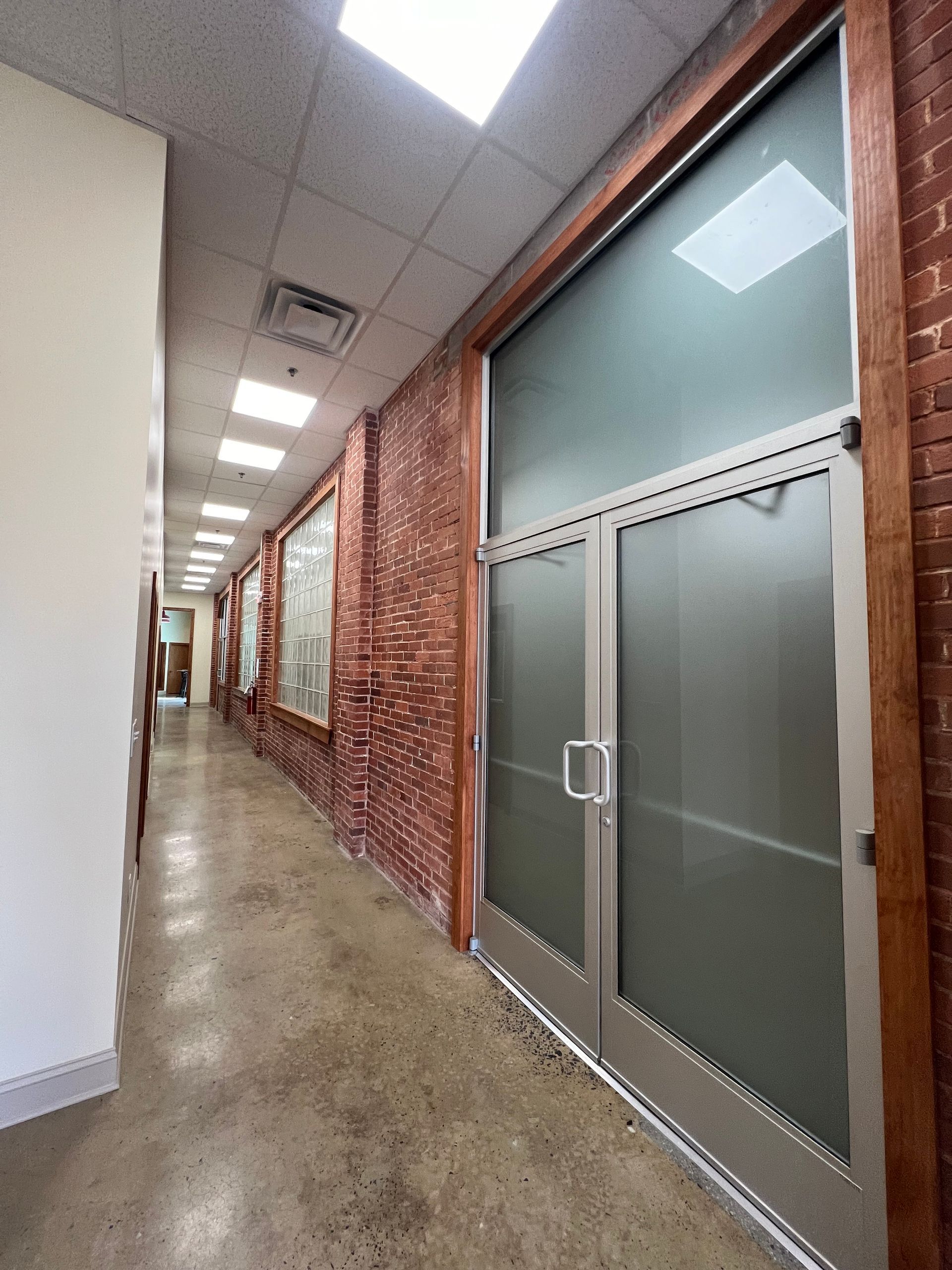 A narrow hallway with a concrete floor, a rustic exposed brick wall, and a set of frosted glass double doors.