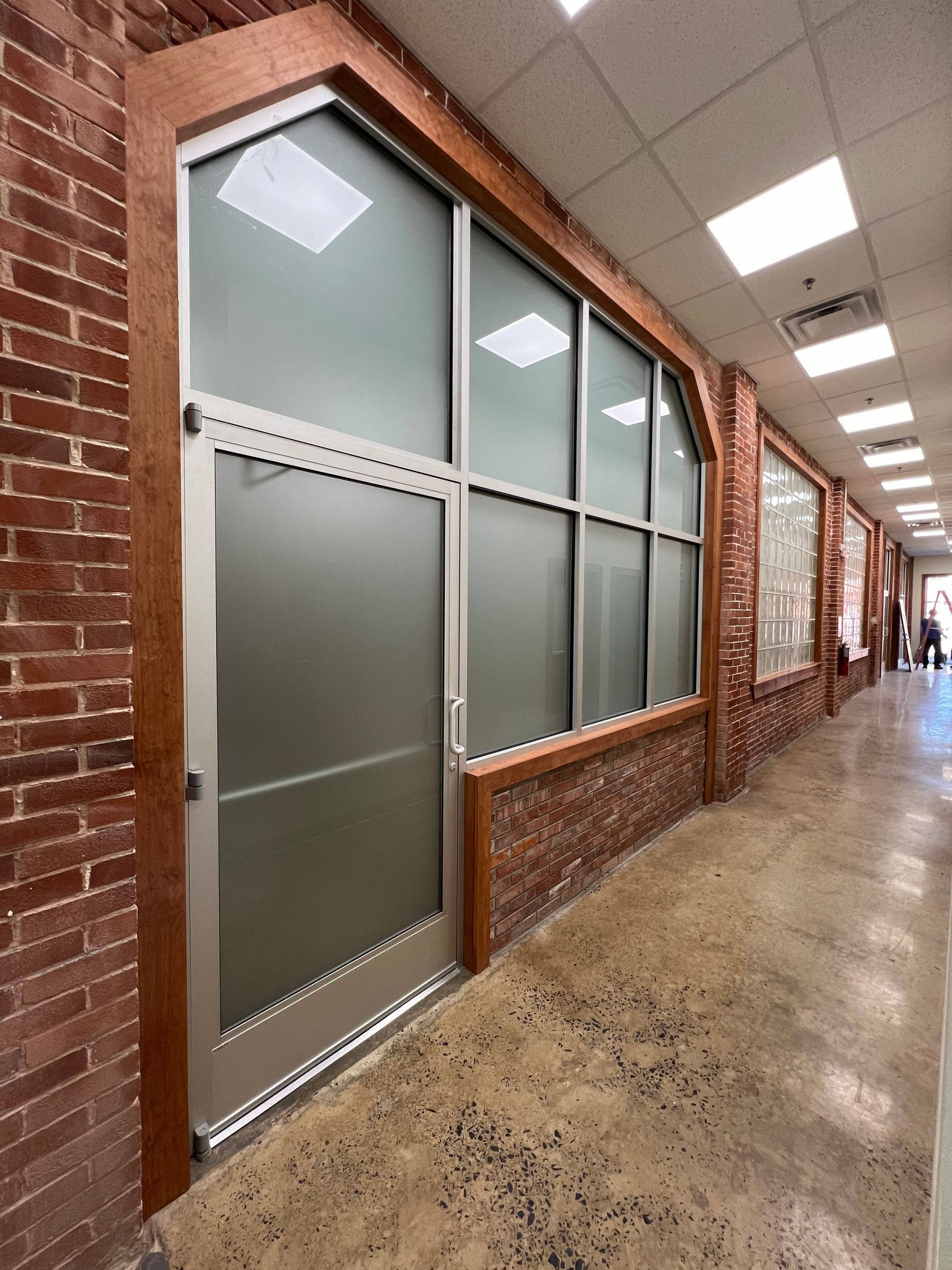 An indoor hallway with exposed brick walls, polished concrete floors, and a frosted glass office entrance with wood trim.