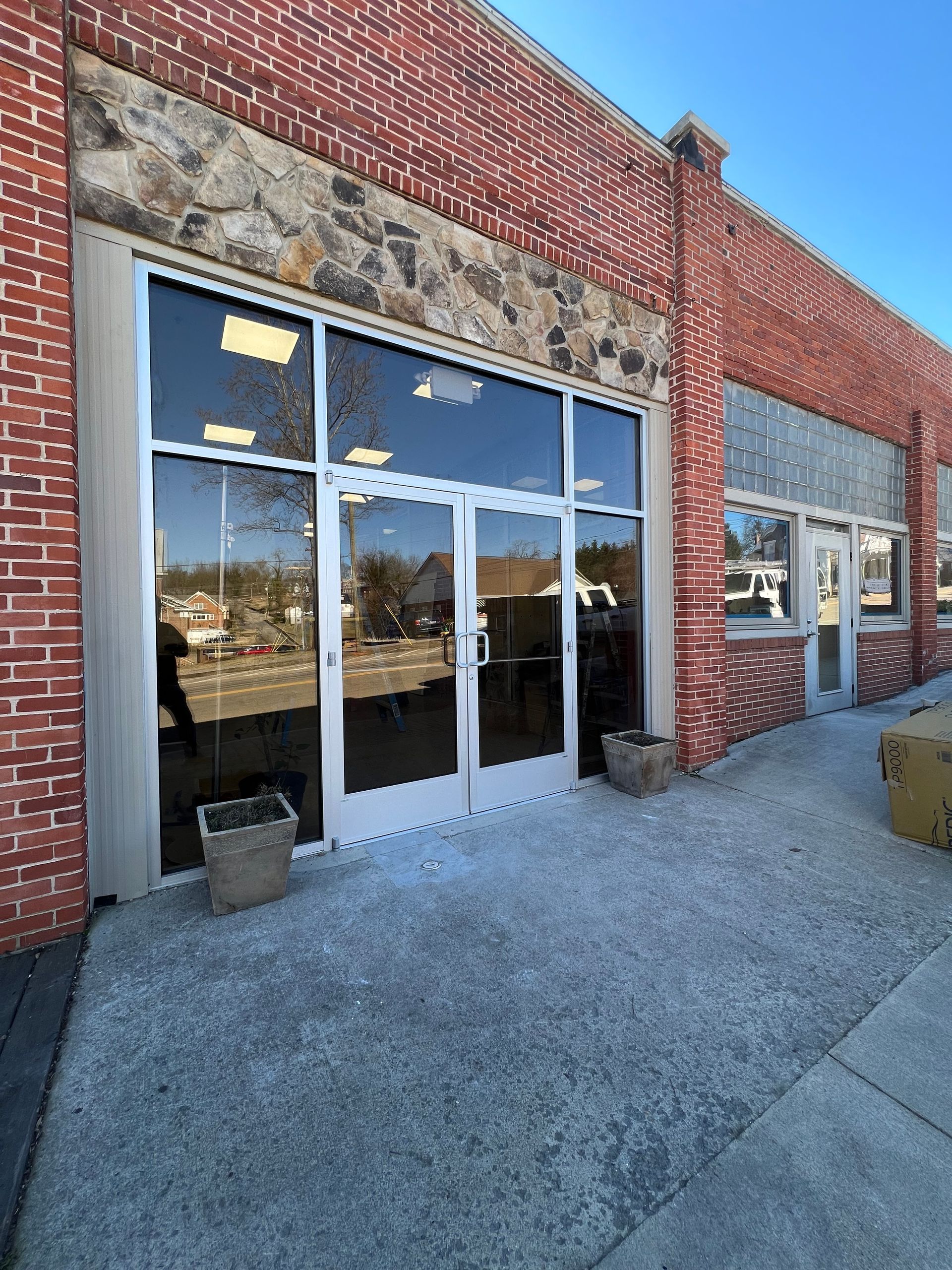 A red brick storefront with large glass double doors and a stone facade, situated on a concrete sidewalk under blue skies.