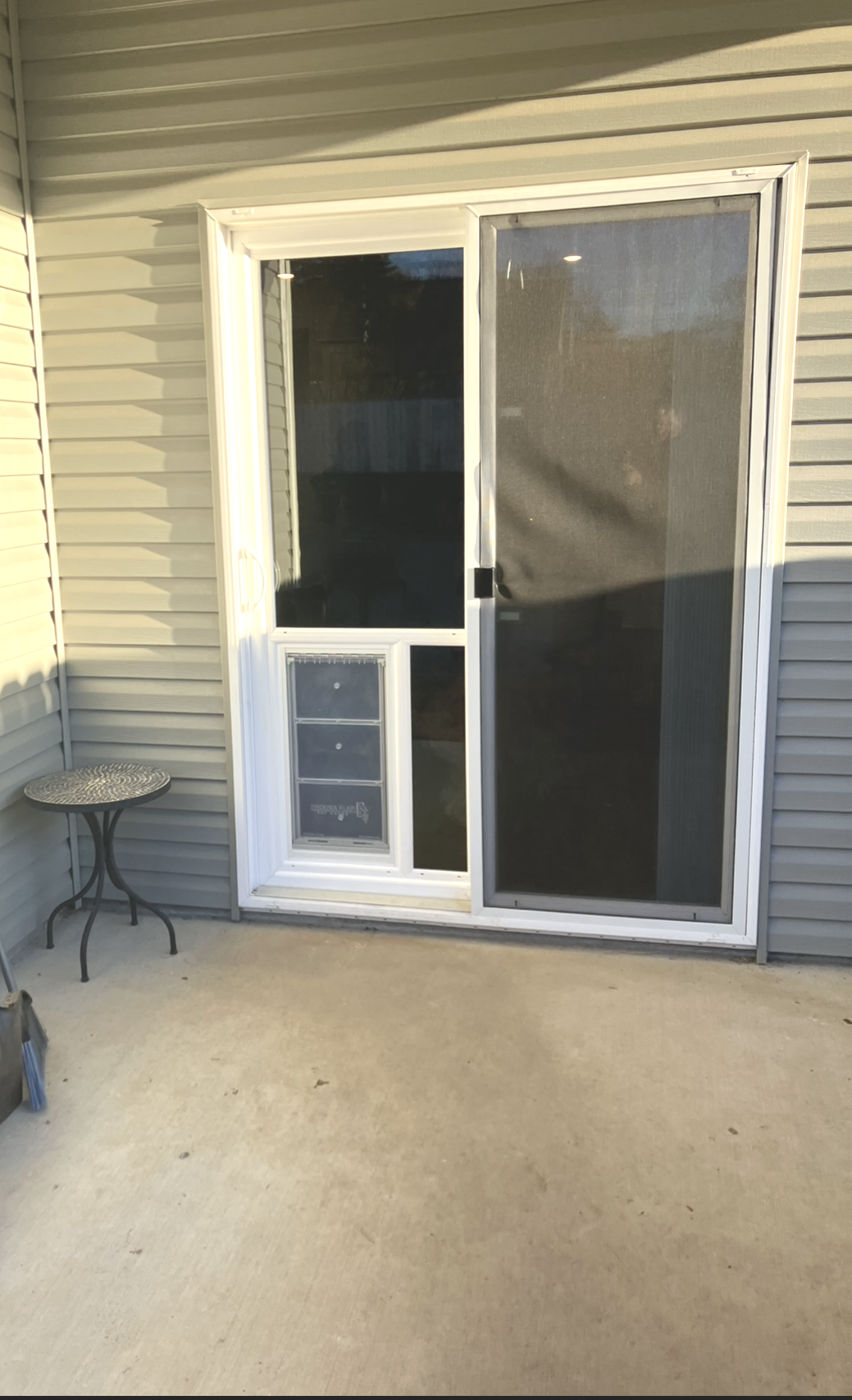 A white sliding glass door with a built-in pet door, set into a beige vinyl-sided exterior wall on a concrete patio.