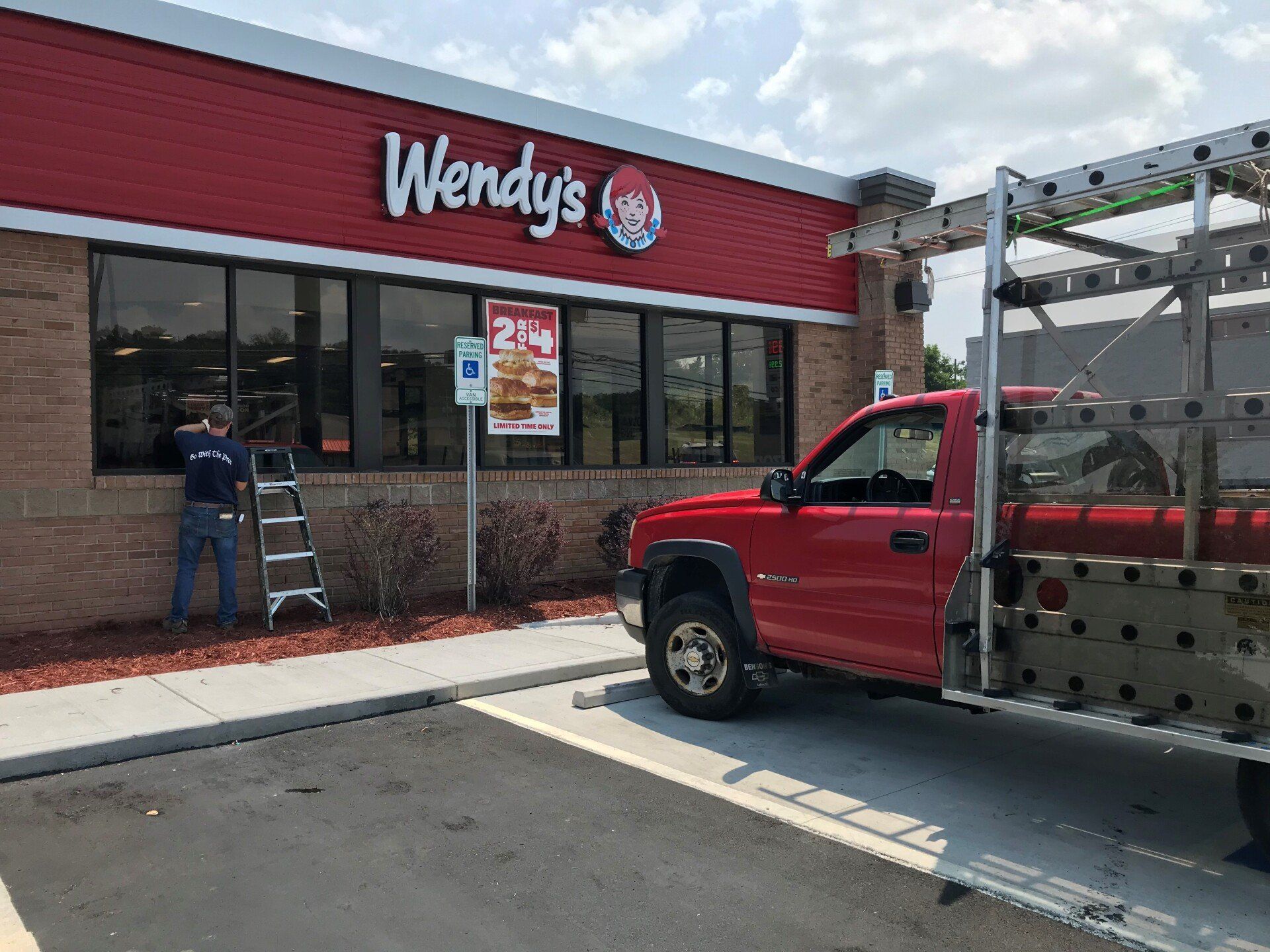 A worker on a ladder installs glass on the exterior of a red-brick Wendy's restaurant next to a utility truck.