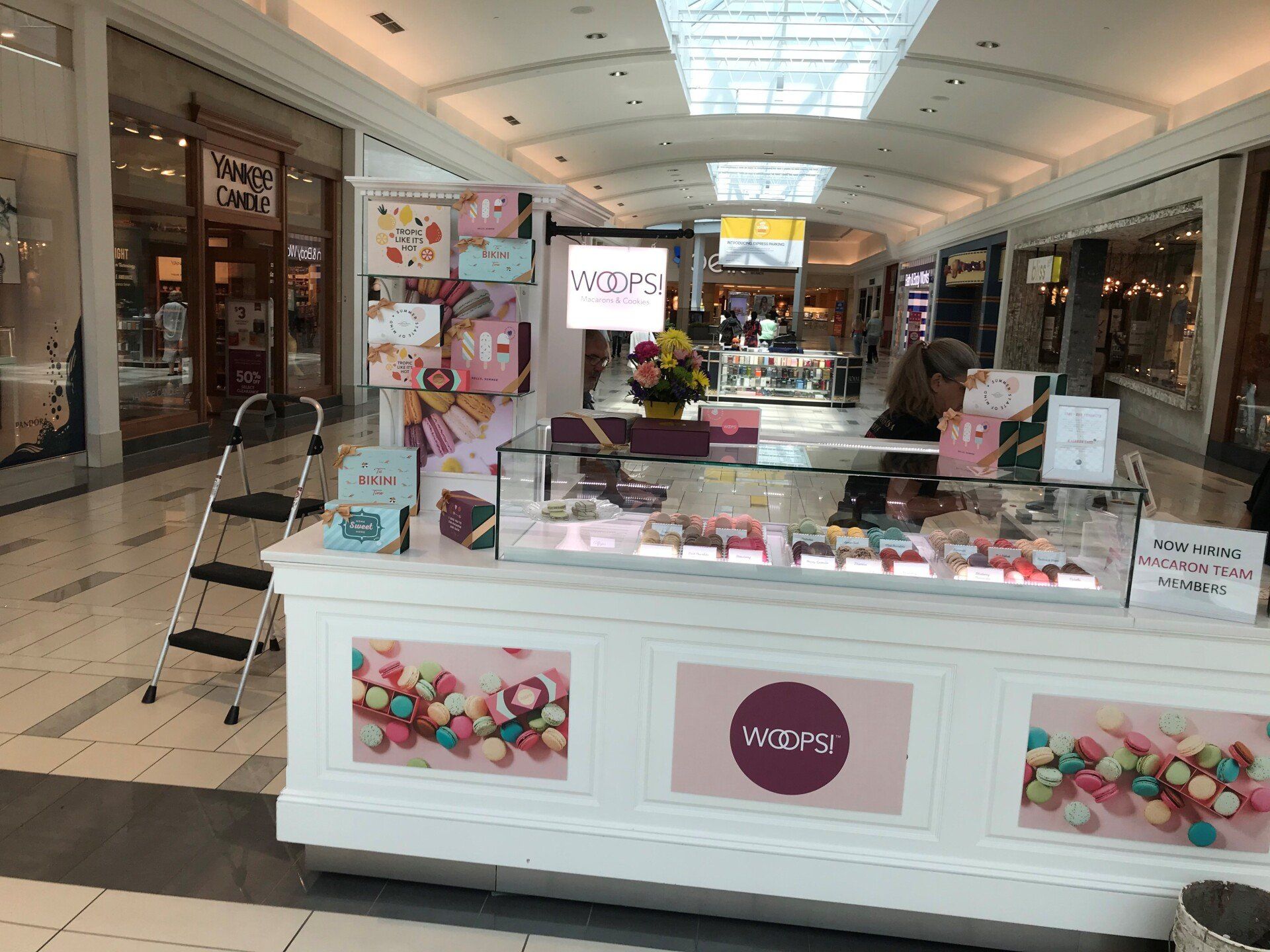 A brightly lit macaron kiosk in a shopping mall, featuring glass display cases and pink branding.