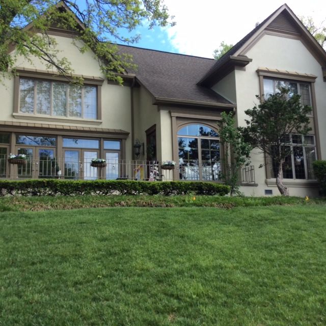 Two-story beige house with brown trim and a large green lawn under a partly cloudy blue sky.