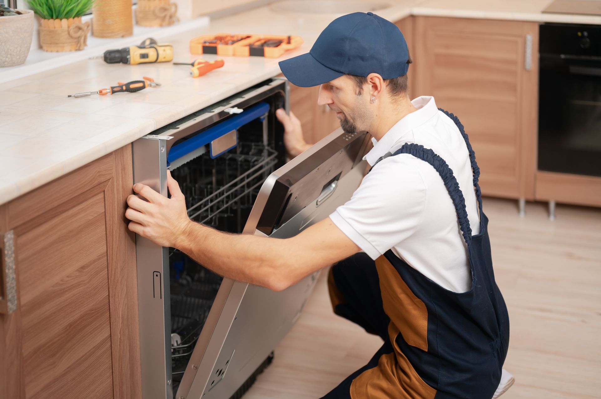 A repairman in a blue cap and overalls, fixing a dishwasher in a kitchen.