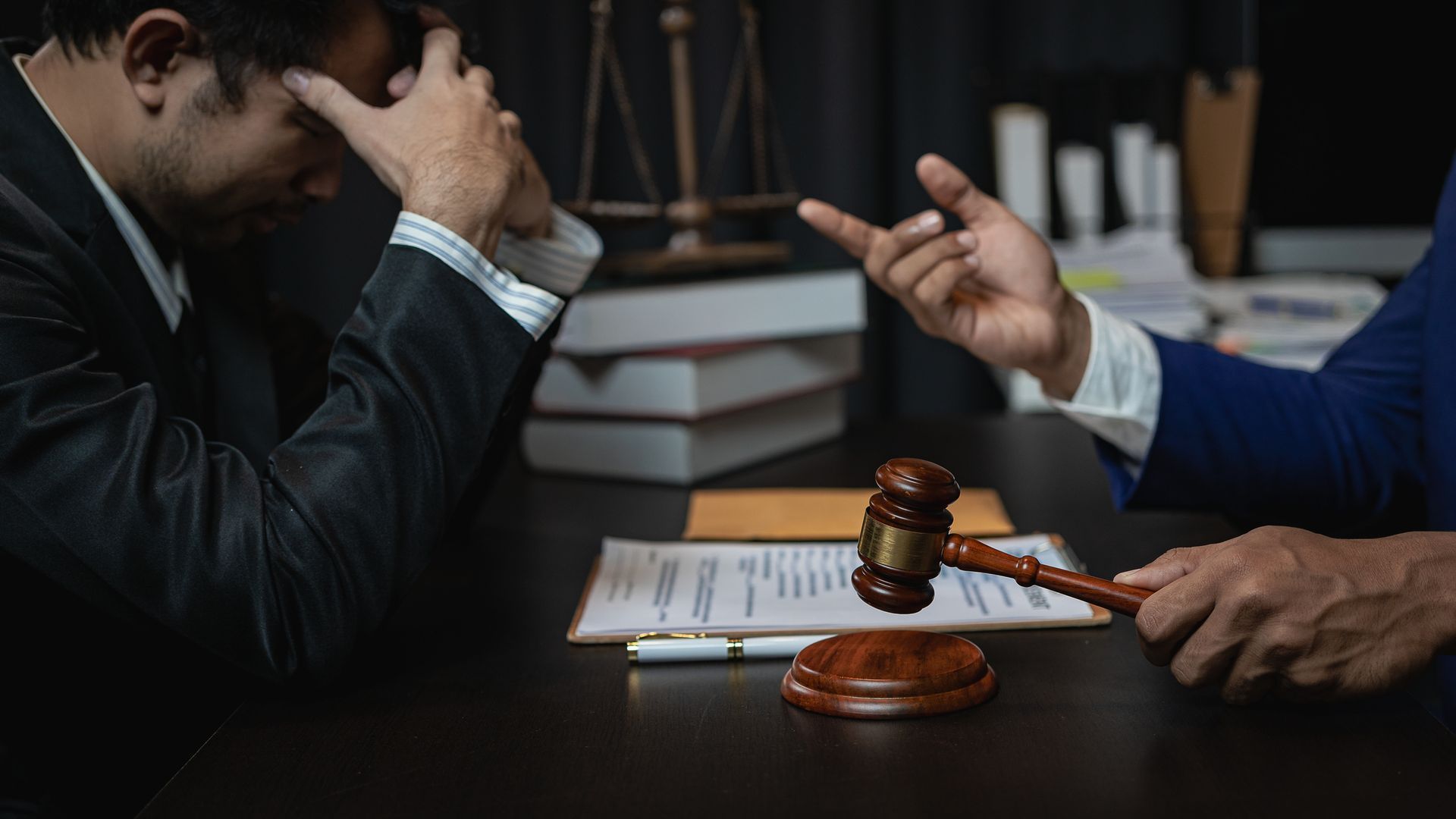 Man in suit looking distressed, opposite a hand holding a gavel at a desk in an office.