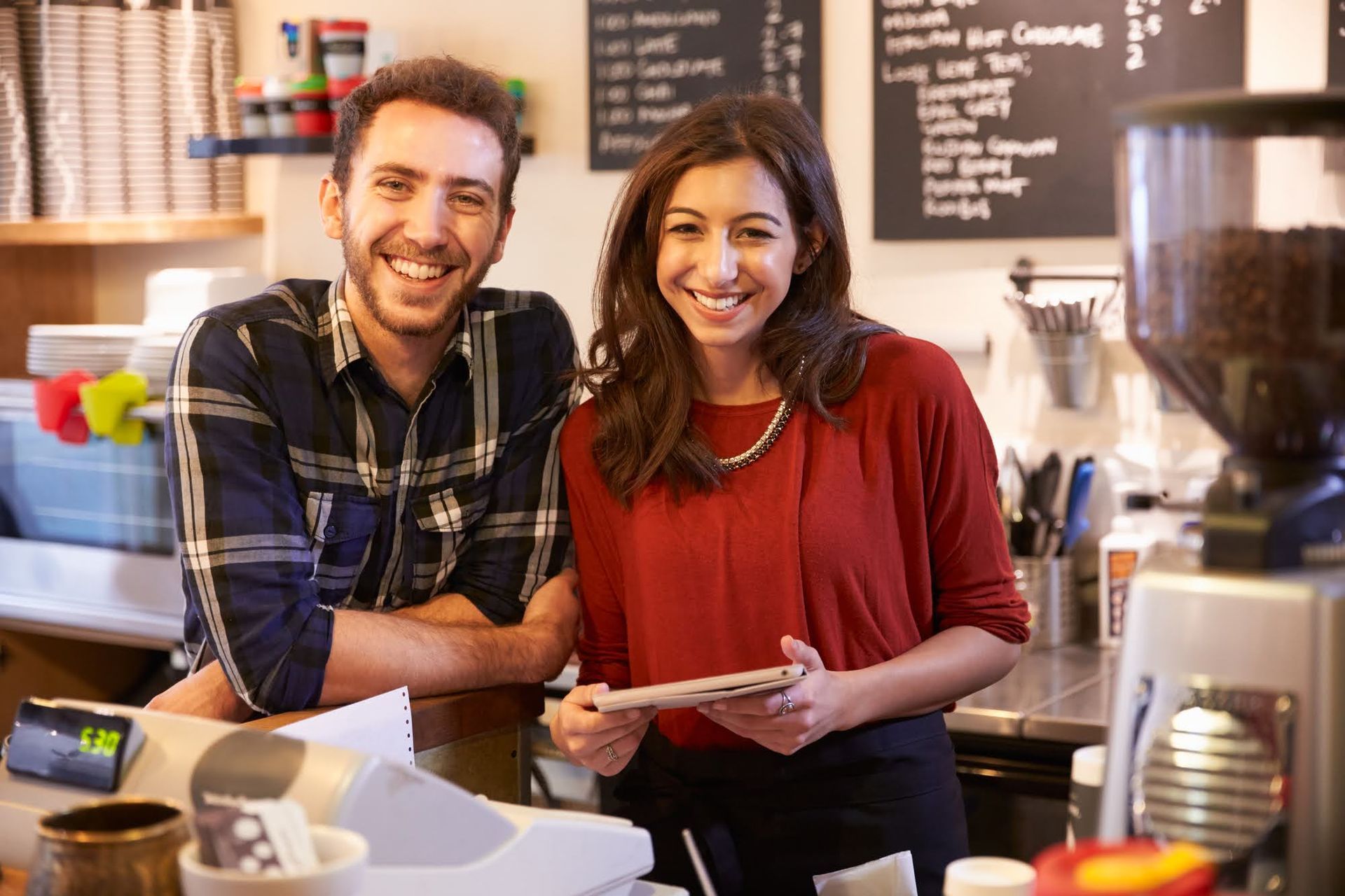 Two people smiling behind a coffee shop counter. Woman holds tablet, man leans on counter.