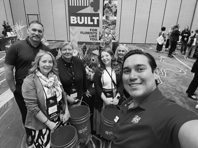 Group selfie at a trade show booth with smiling attendees holding branded buckets.