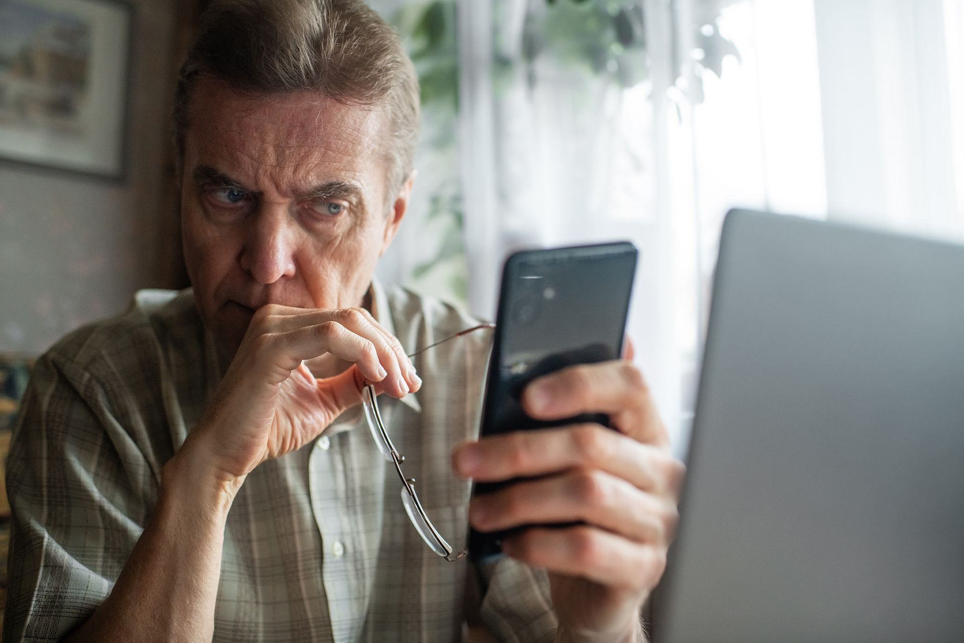 An older man is looking at his cell phone while sitting in front of a laptop.