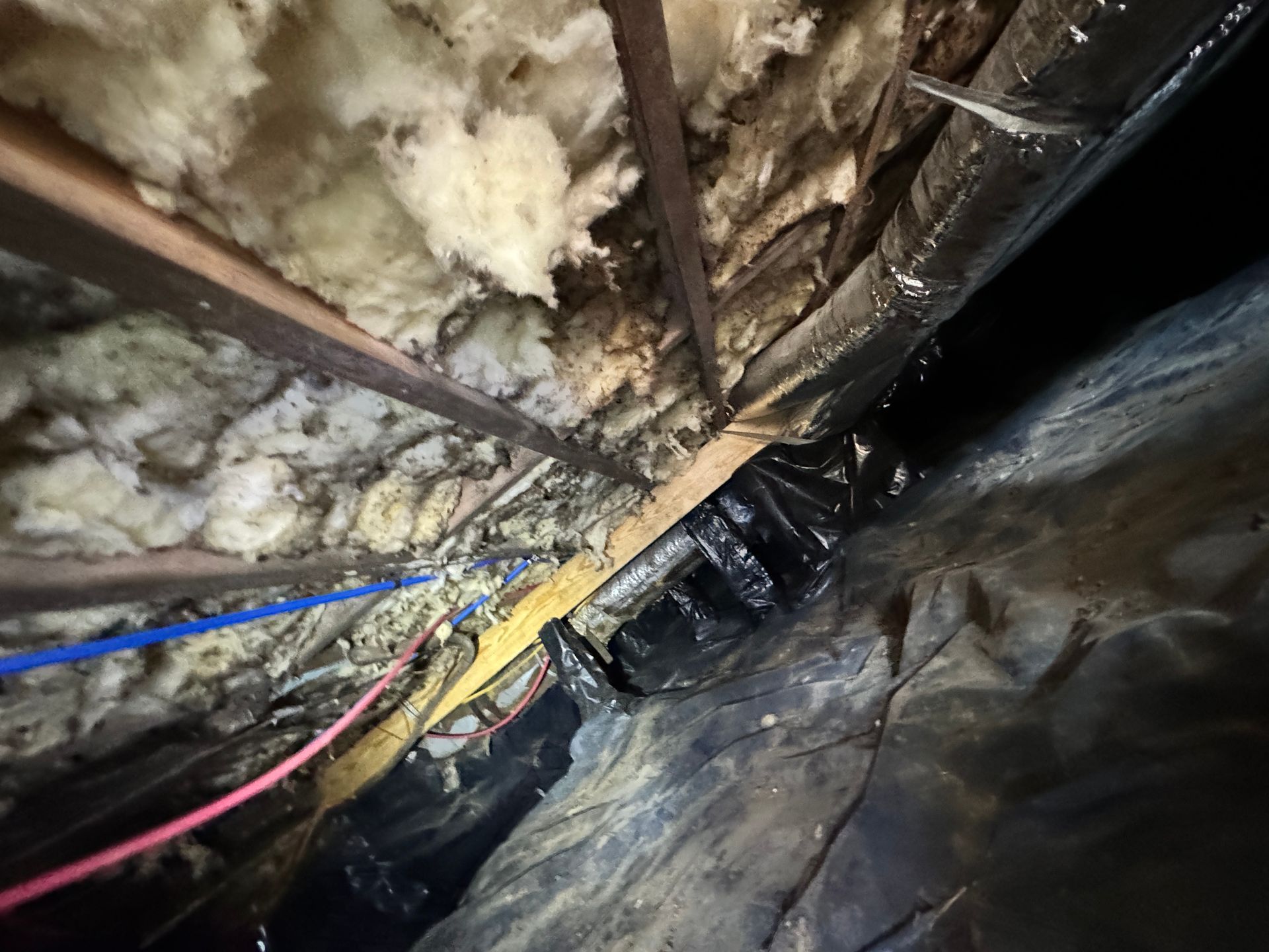 View of an attic with insulation, beams, and black plastic sheeting. Electrical wiring and pipes are visible.