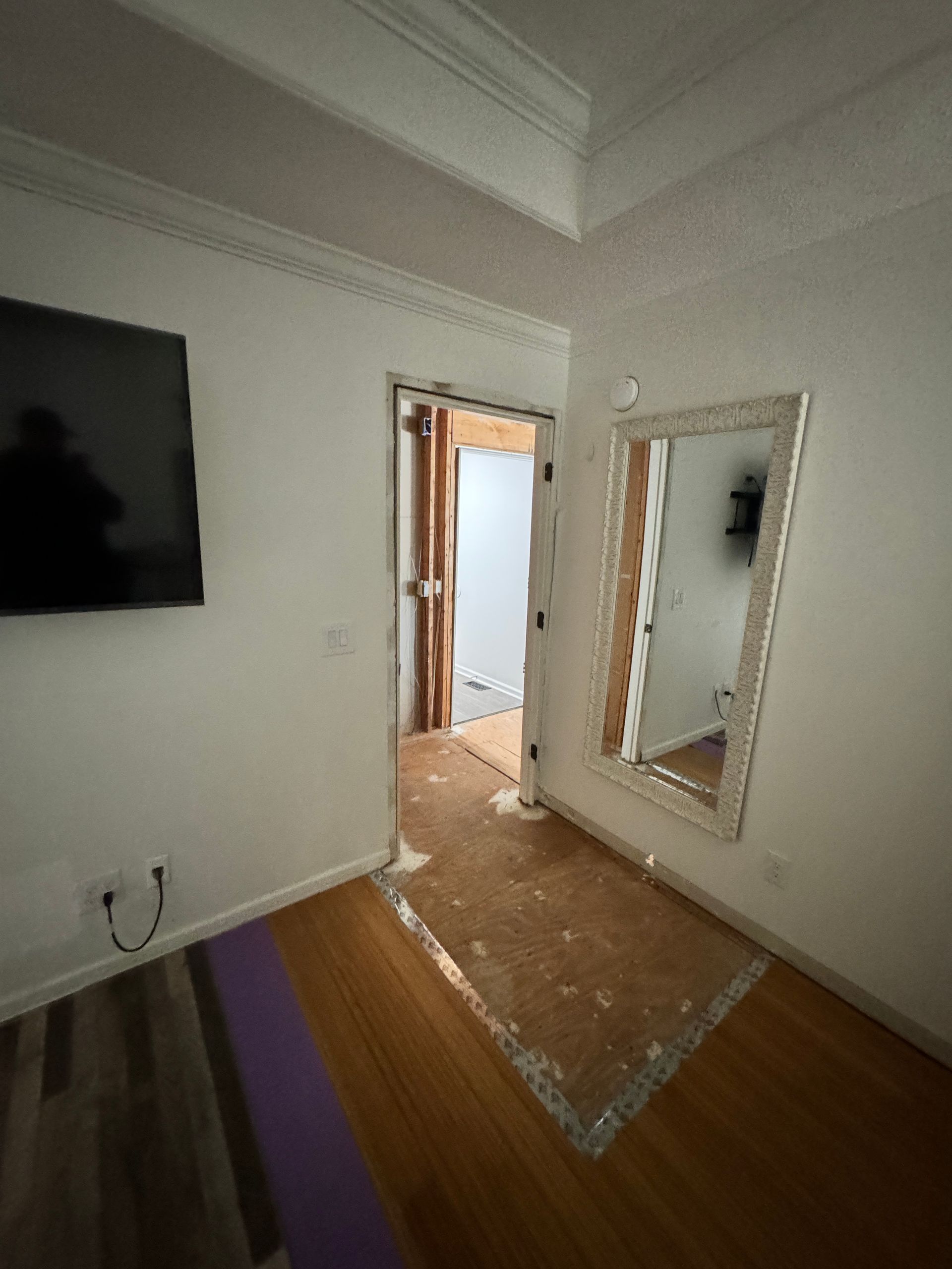 Bedroom interior with hardwood floor, doorway, mirror, TV, white walls, and partial demolition.