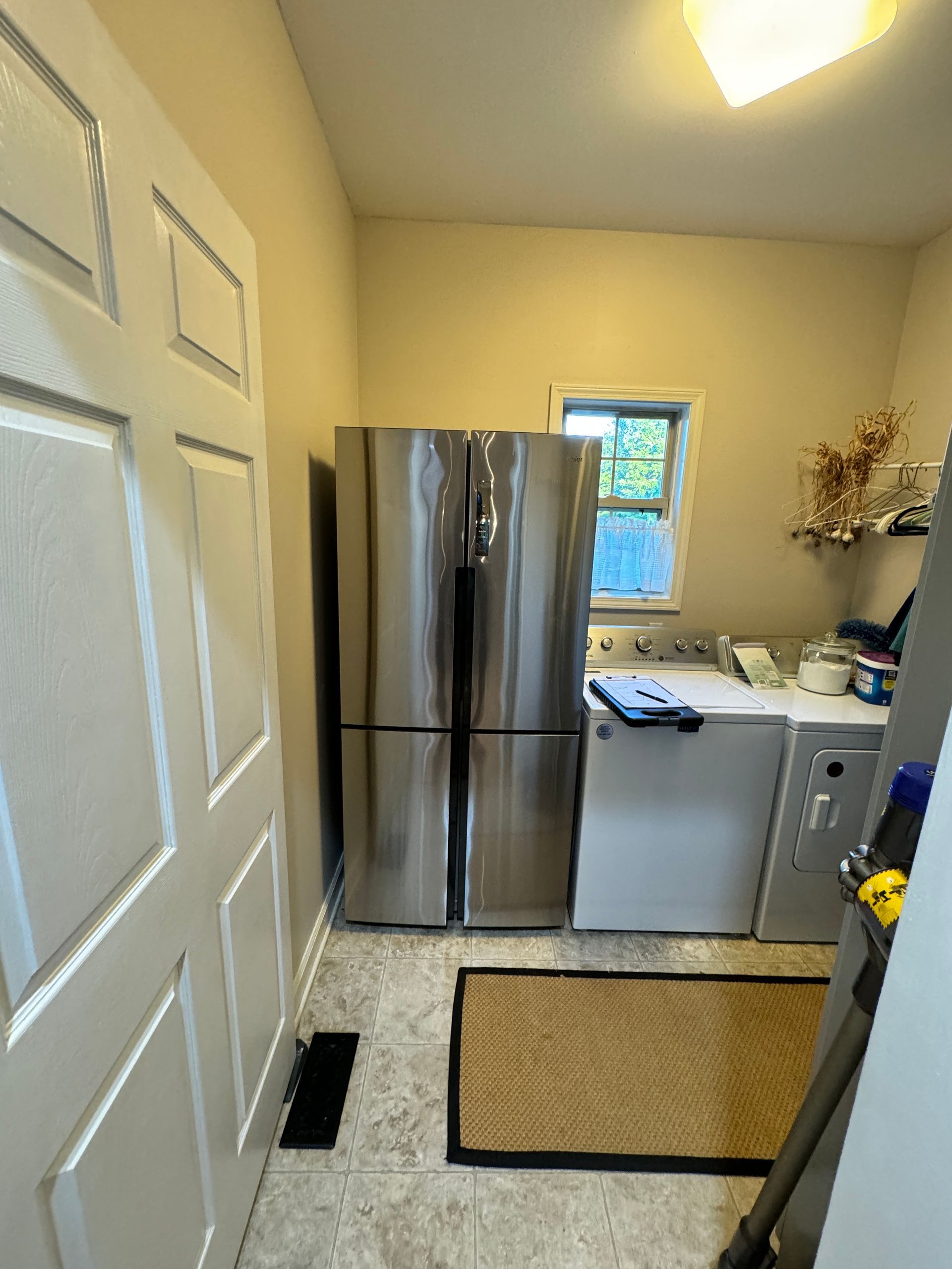 Laundry room with stainless steel fridge, washer, dryer, door, and small window. Beige walls, light brown mat.