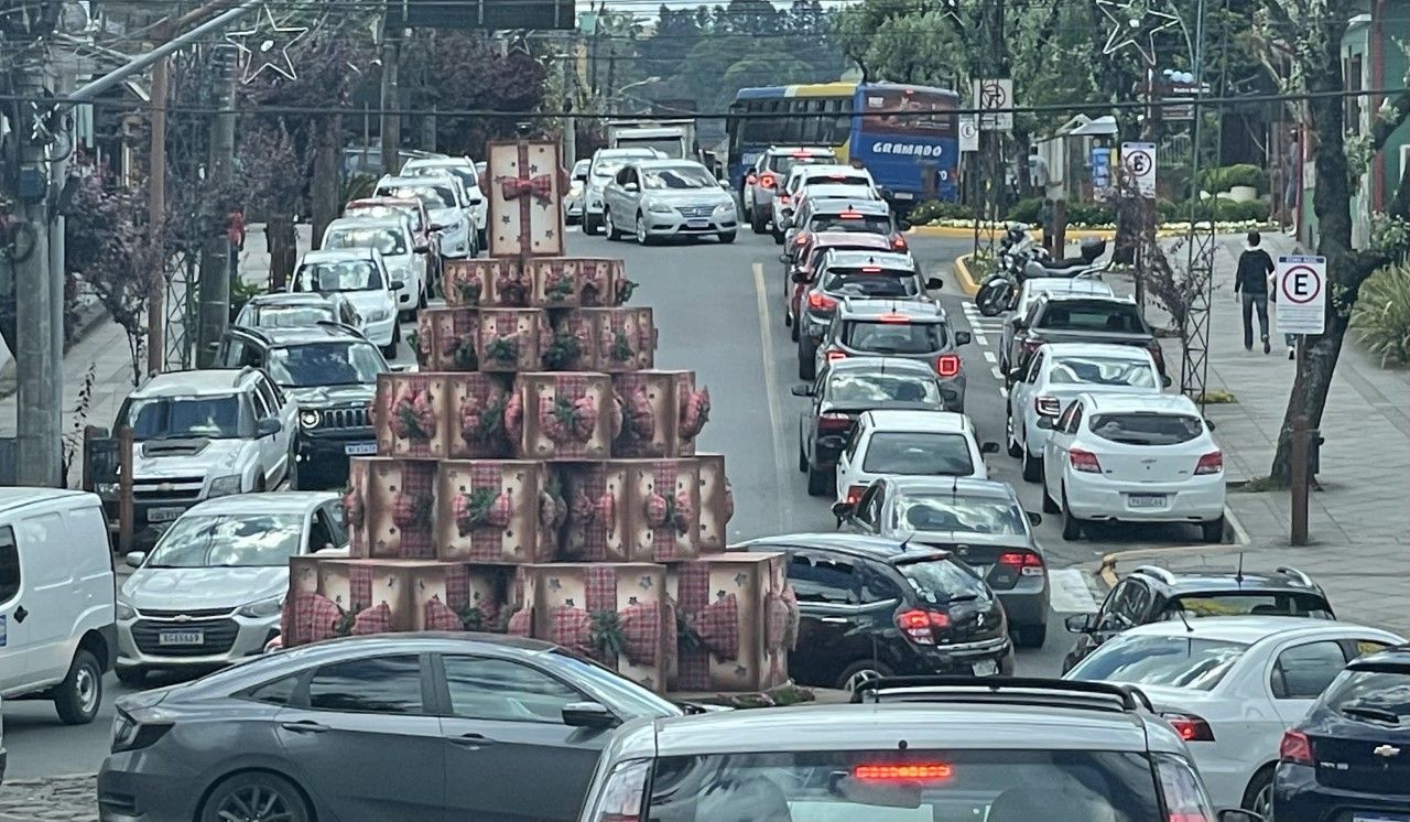 Muitos carros passam por uma rua com uma árvore de natal feita de latas.