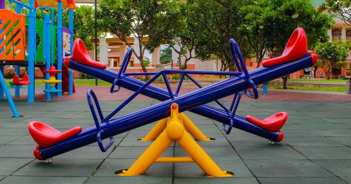A very colorful playground with see-saws, a climbing structure, and permeable paving is backed by trees.
